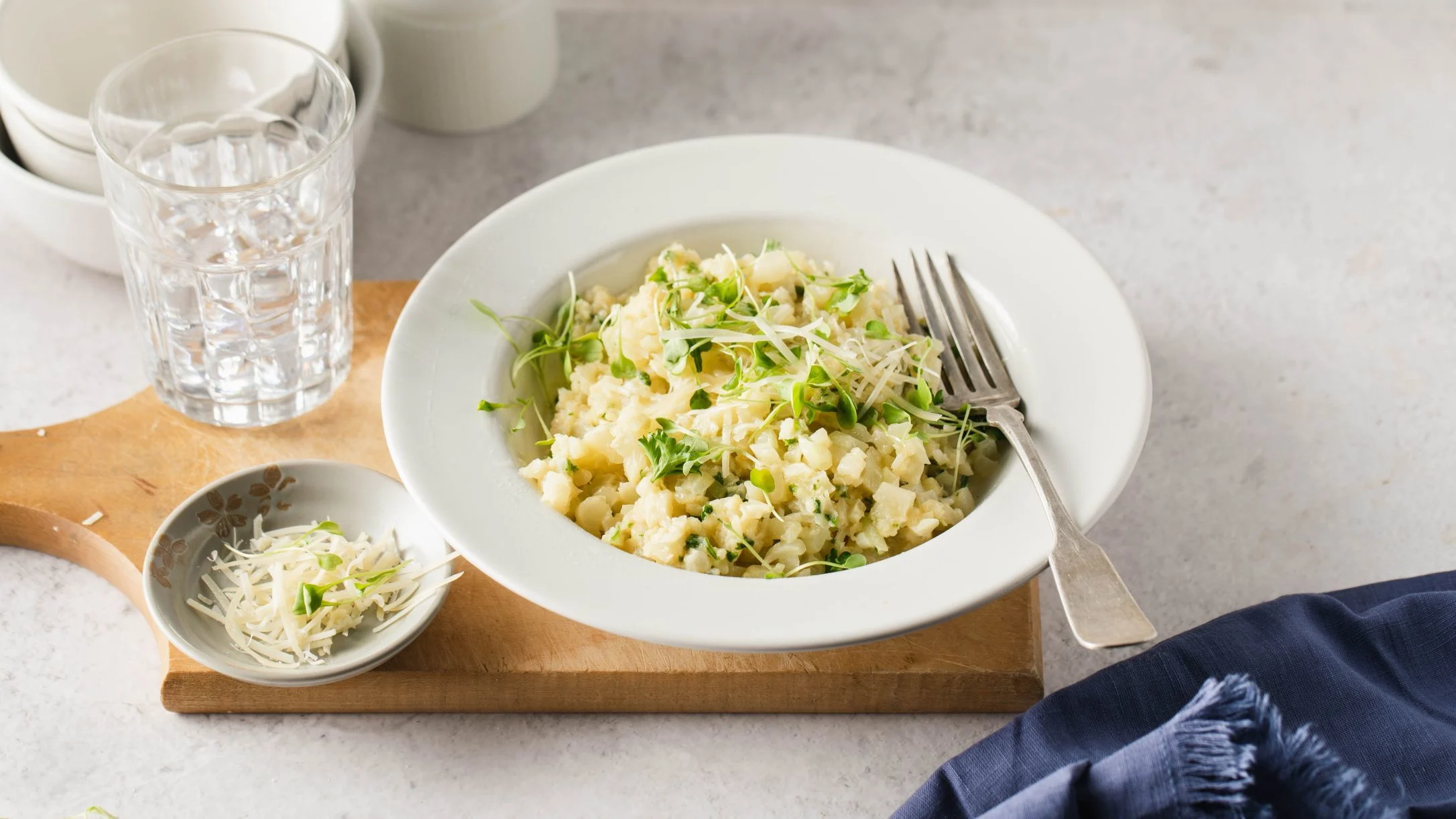 Creamy risotto topped with microgreens served in white bowl with fork, glass of water, and small dish of garnish nearby.