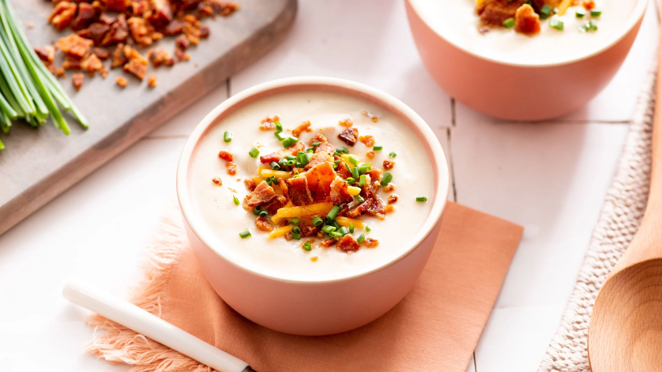 Creamy faux potato soup in pink bowls topped with crispy bacon bits, cheese, and chopped green onions on peach napkins.