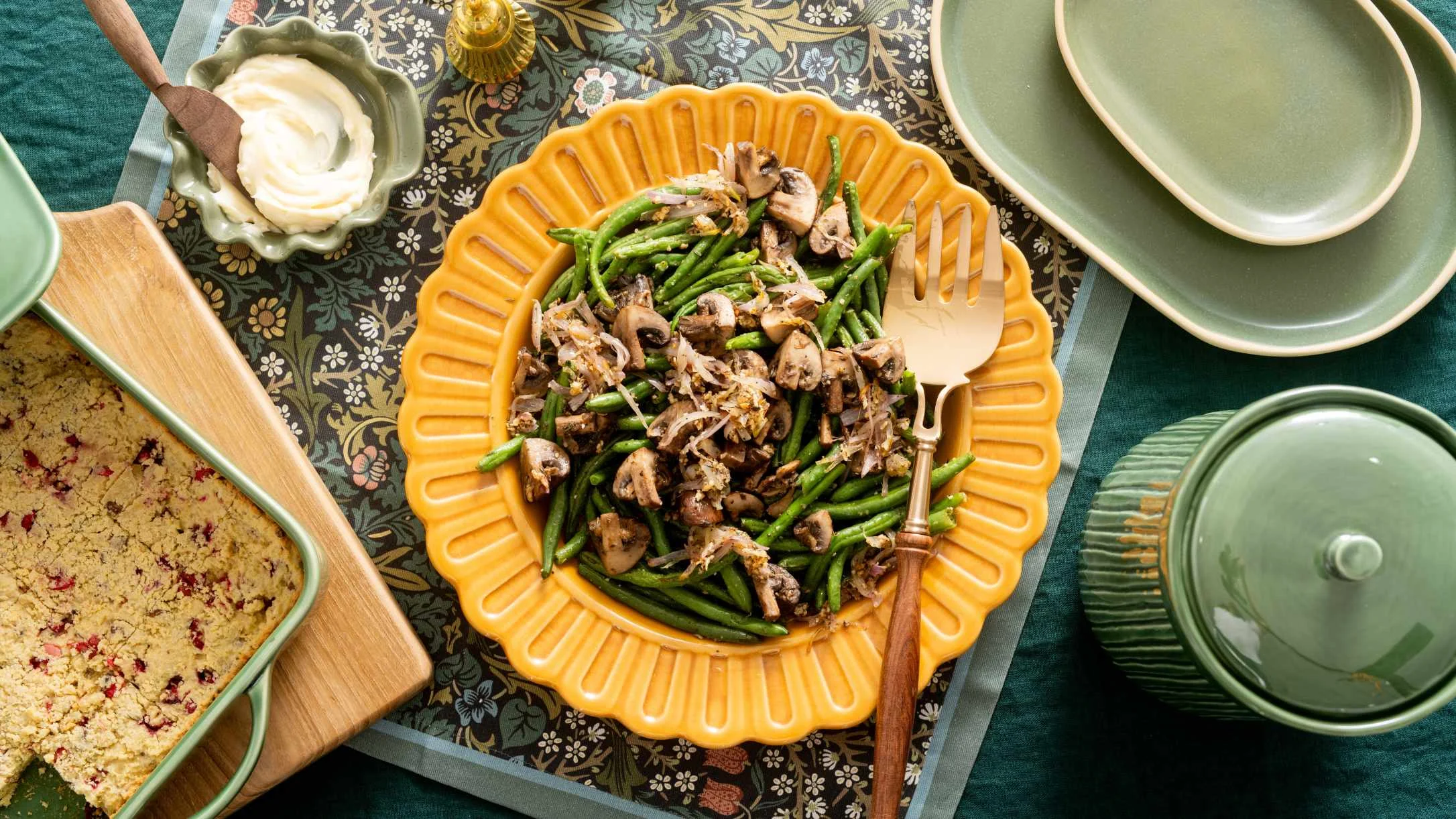 Sautéed mushrooms and green beans in yellow scalloped dish, surrounded by green dinnerware and cream sauce on floral tablecloth.