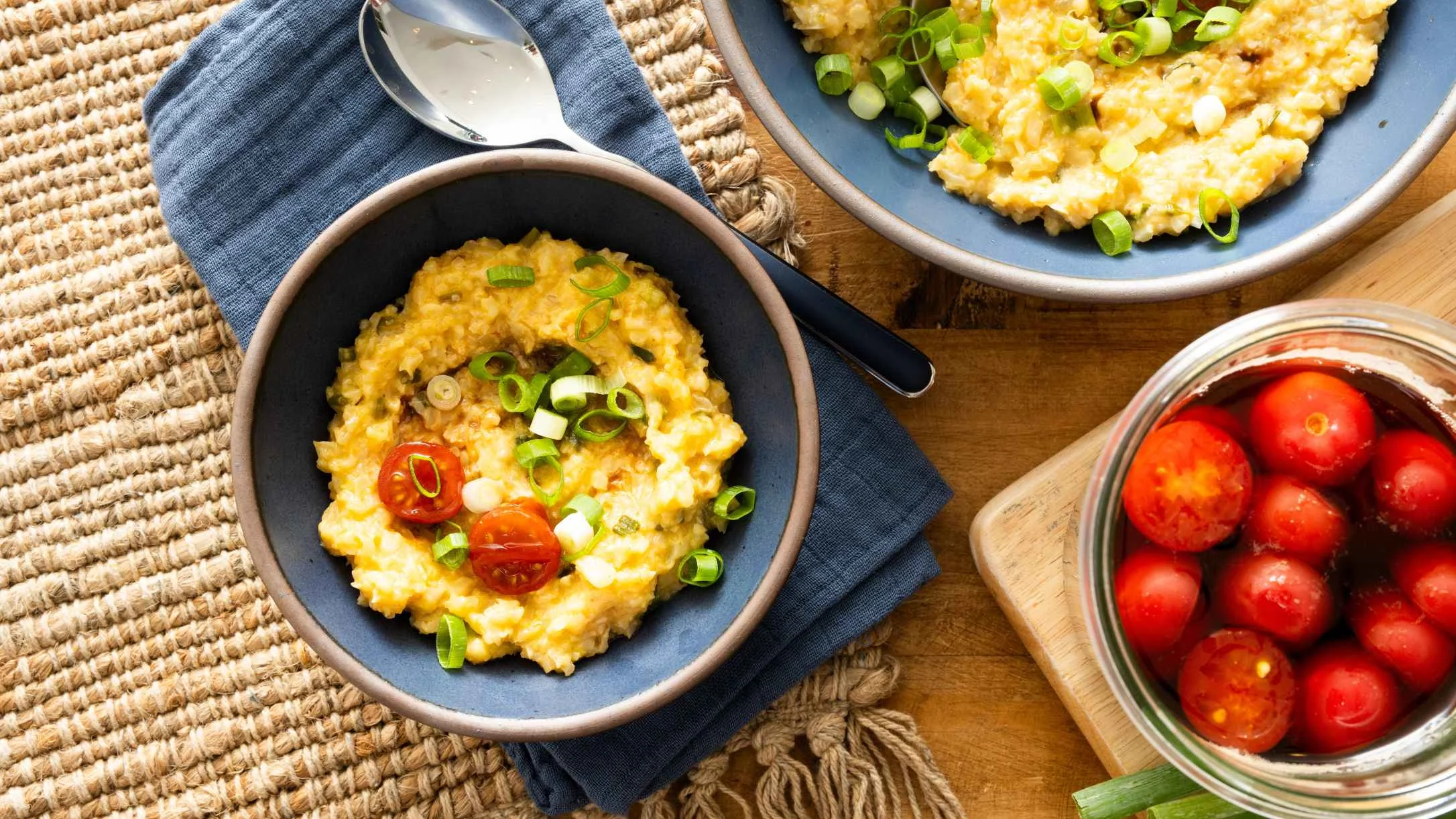 Creamy yellow risotto in blue bowls garnished with green onions and cherry tomatoes, with a bowl of marinating tomatoes nearby.