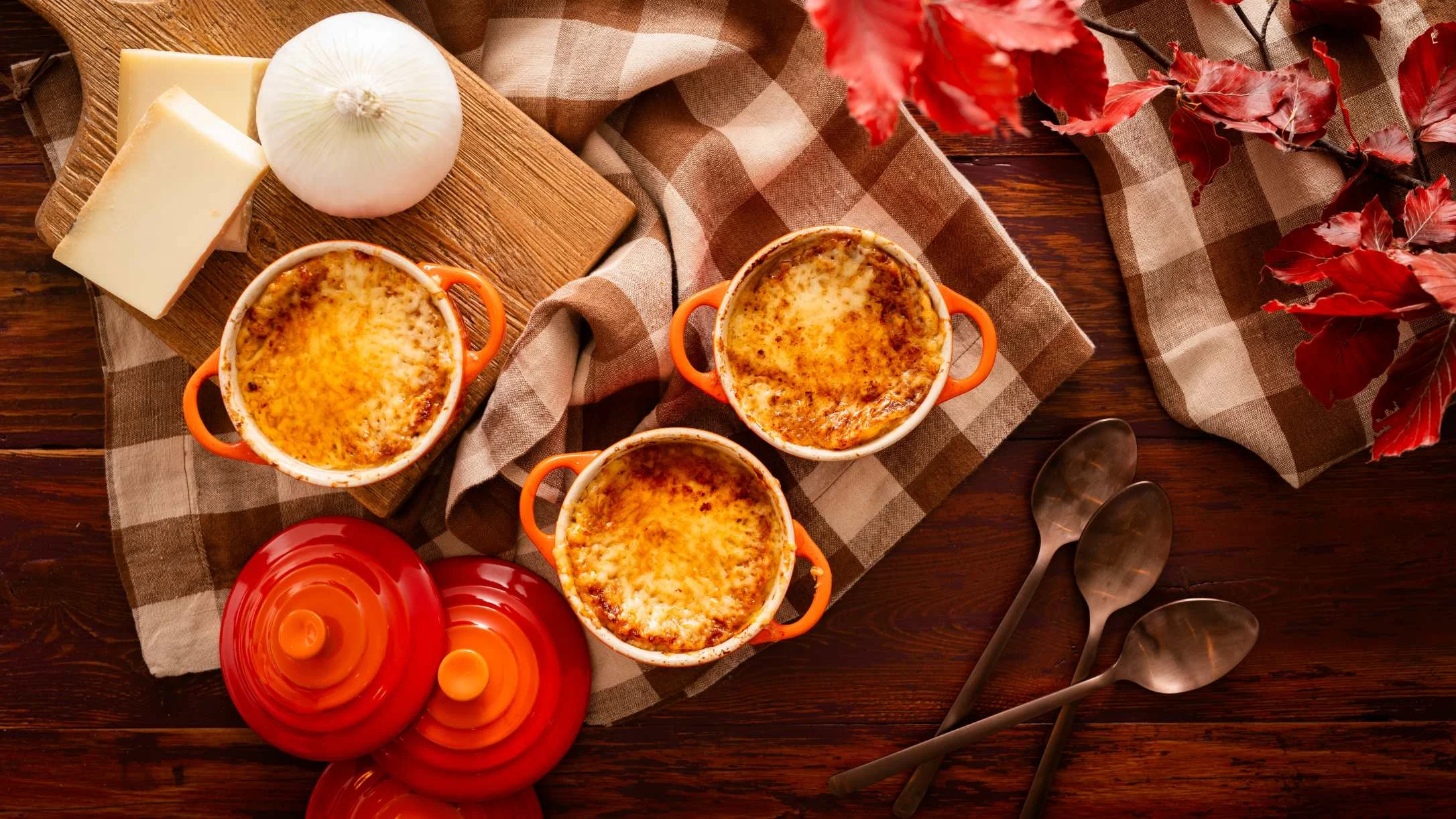 Three orange ramekins of French onion soup with melted cheese on a checkered cloth with cheese, onion, and autumn leaves.