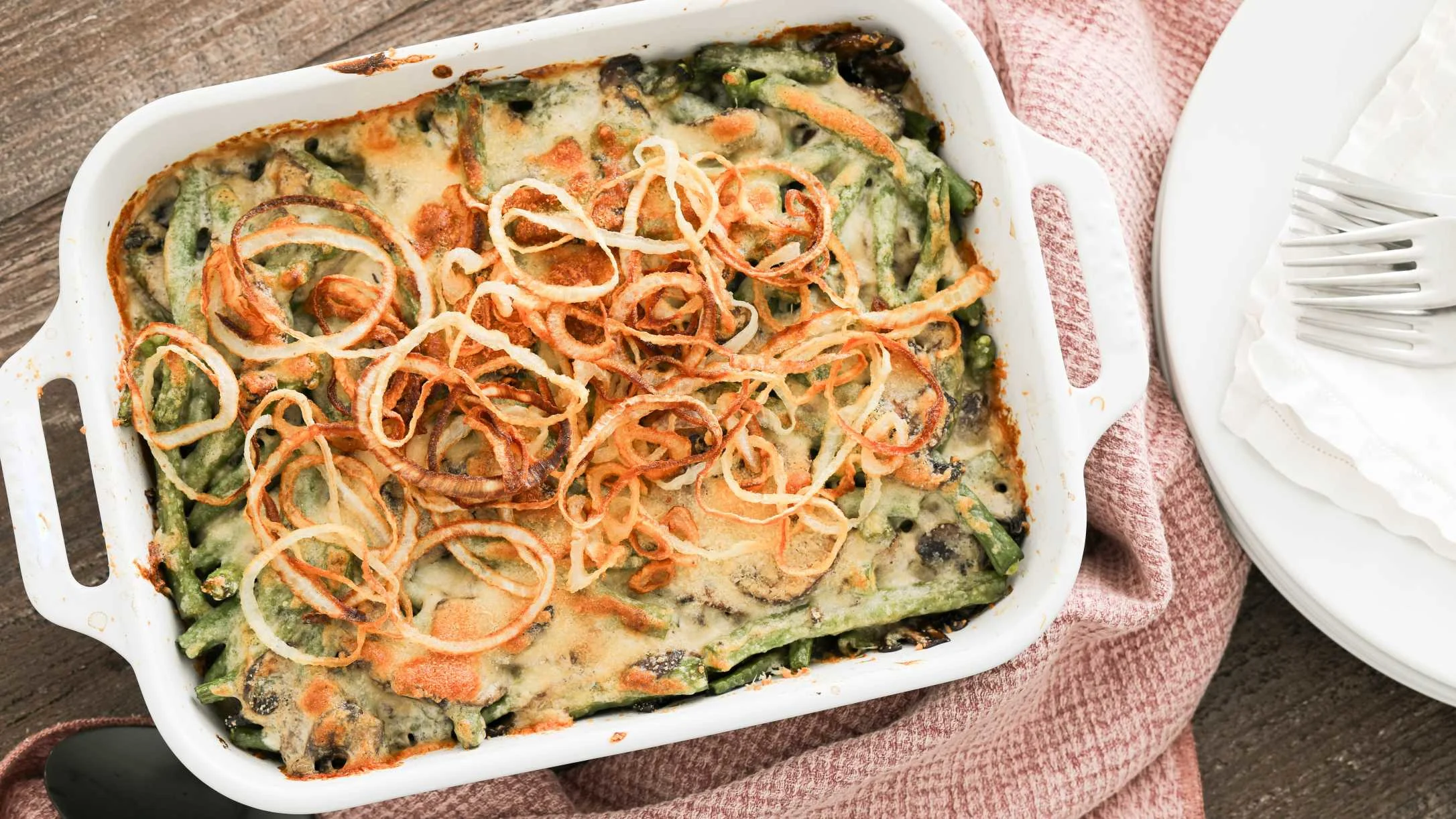 Green bean casserole in white baking dish topped with crispy fried onions, served on pink cloth napkin.