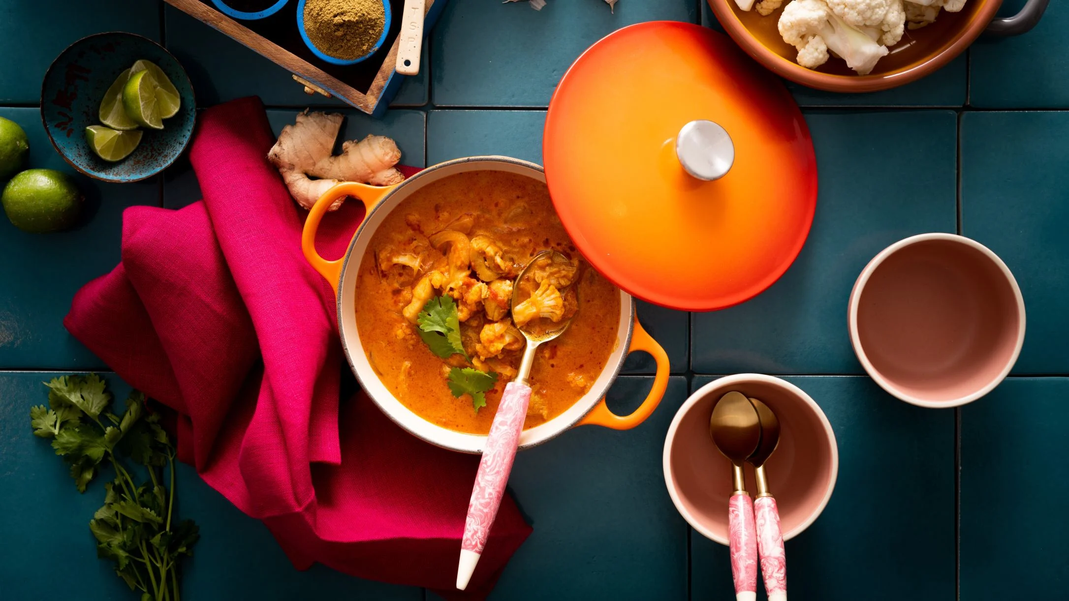 Orange pot of cauliflower curry with lime wedges, ginger, spices, and pink utensils on teal tile surface.