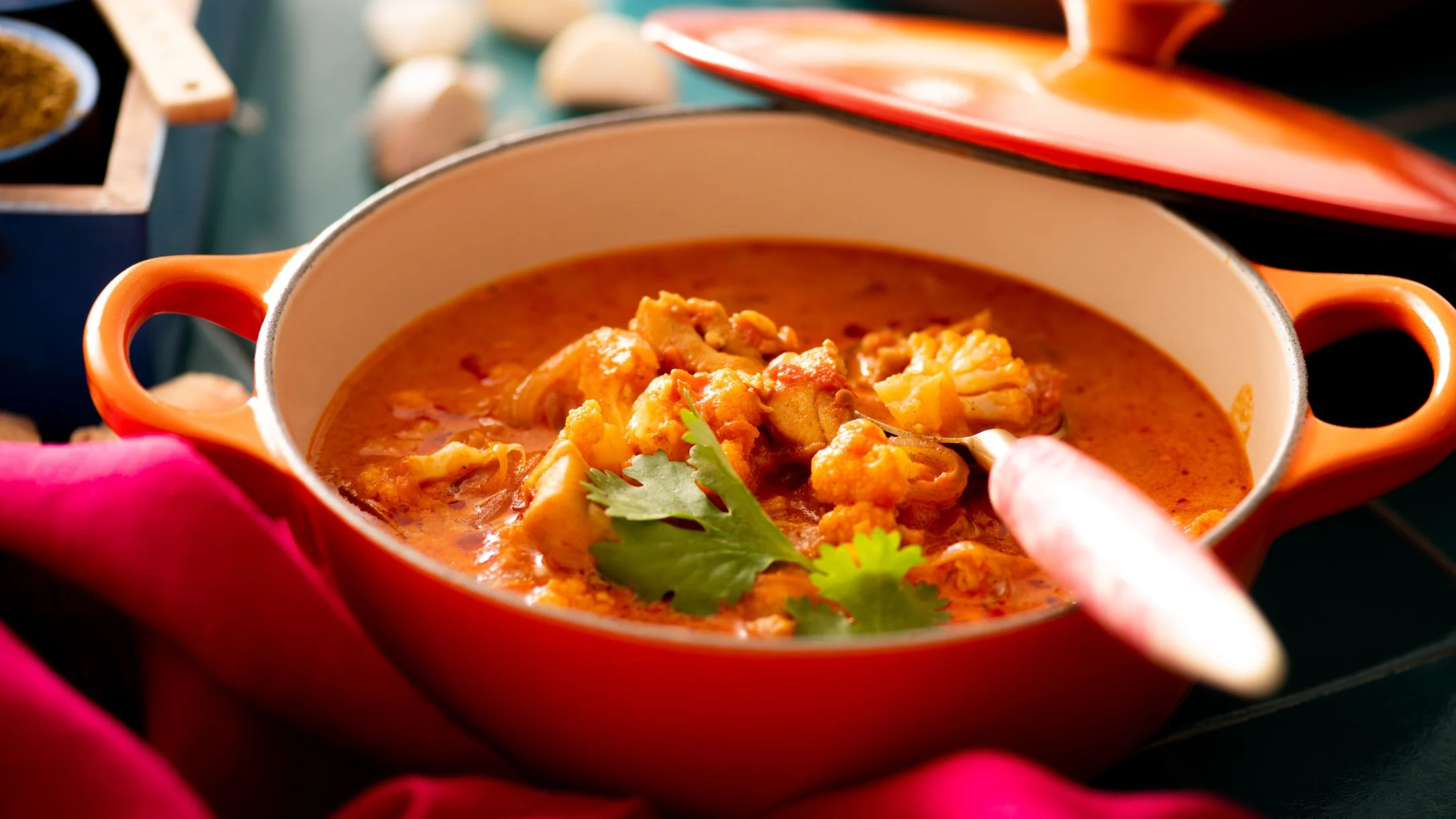 Orange Dutch oven containing spicy curry with cauliflower and chicken, garnished with fresh cilantro leaves.