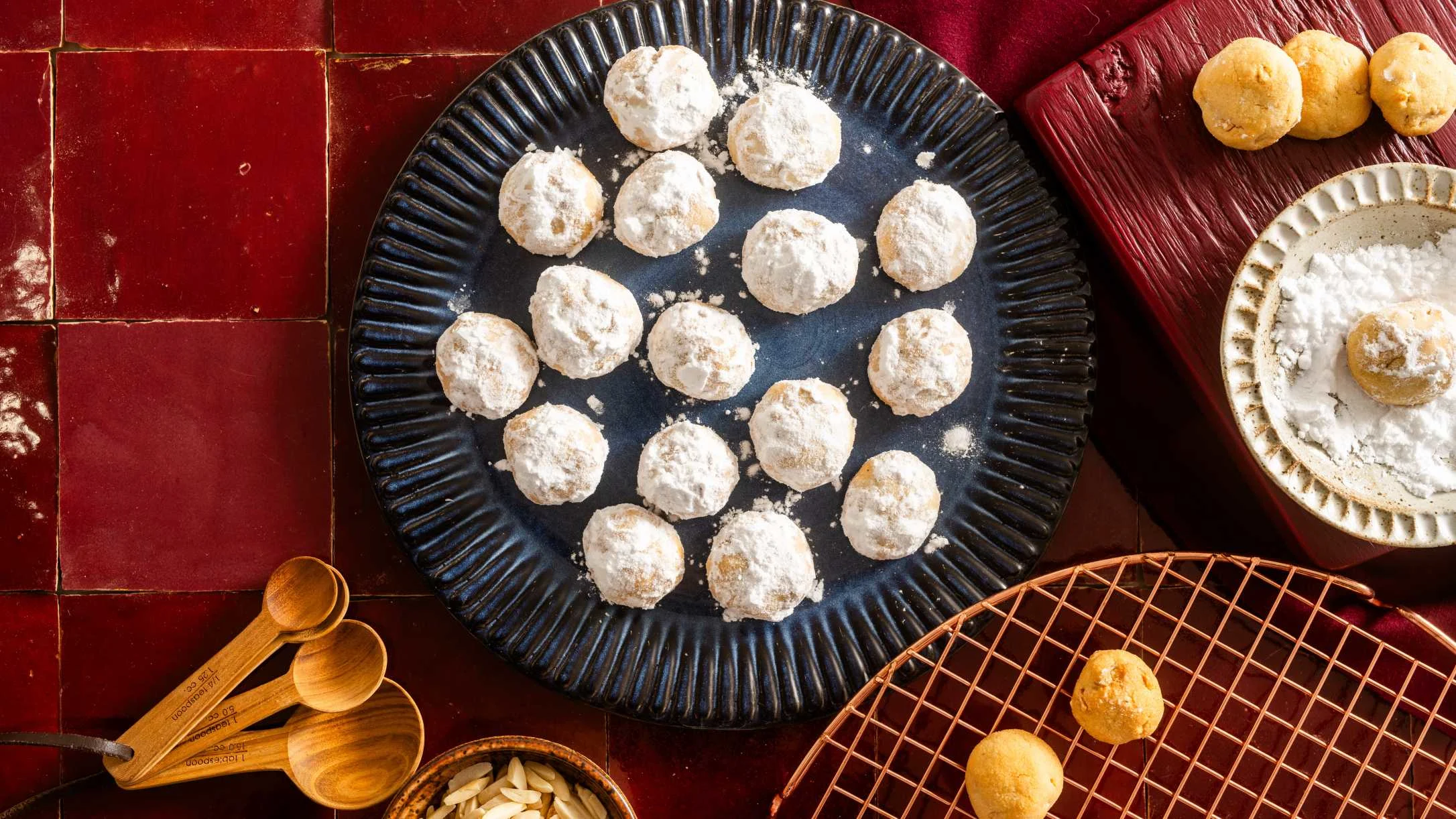 Powdered snowball cookies on a blue plate with baking ingredients and wooden spoons on a red tile surface.