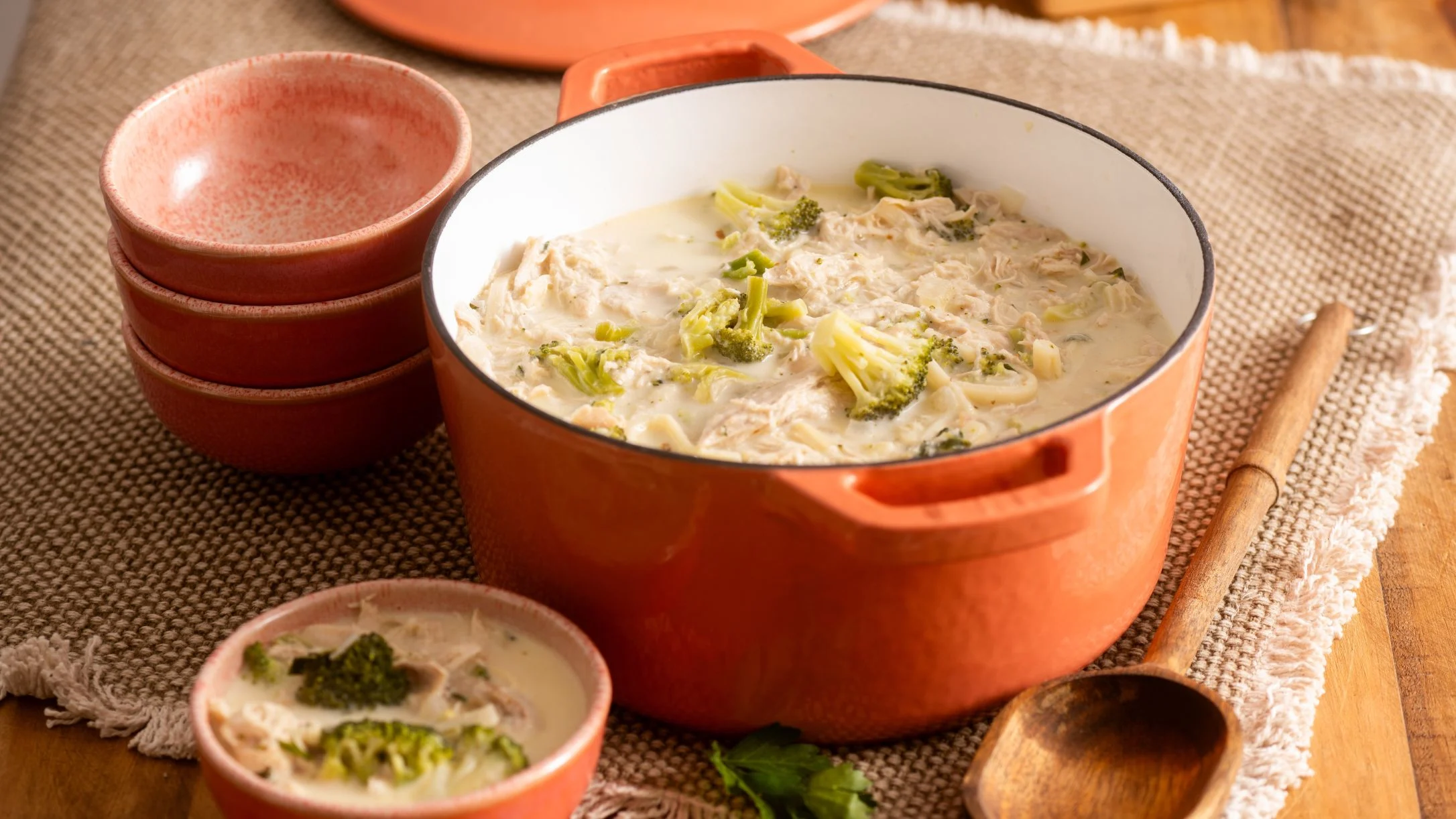 Creamy chicken and broccoli soup in orange pot with matching bowls on textured placemat, wooden spoon nearby.