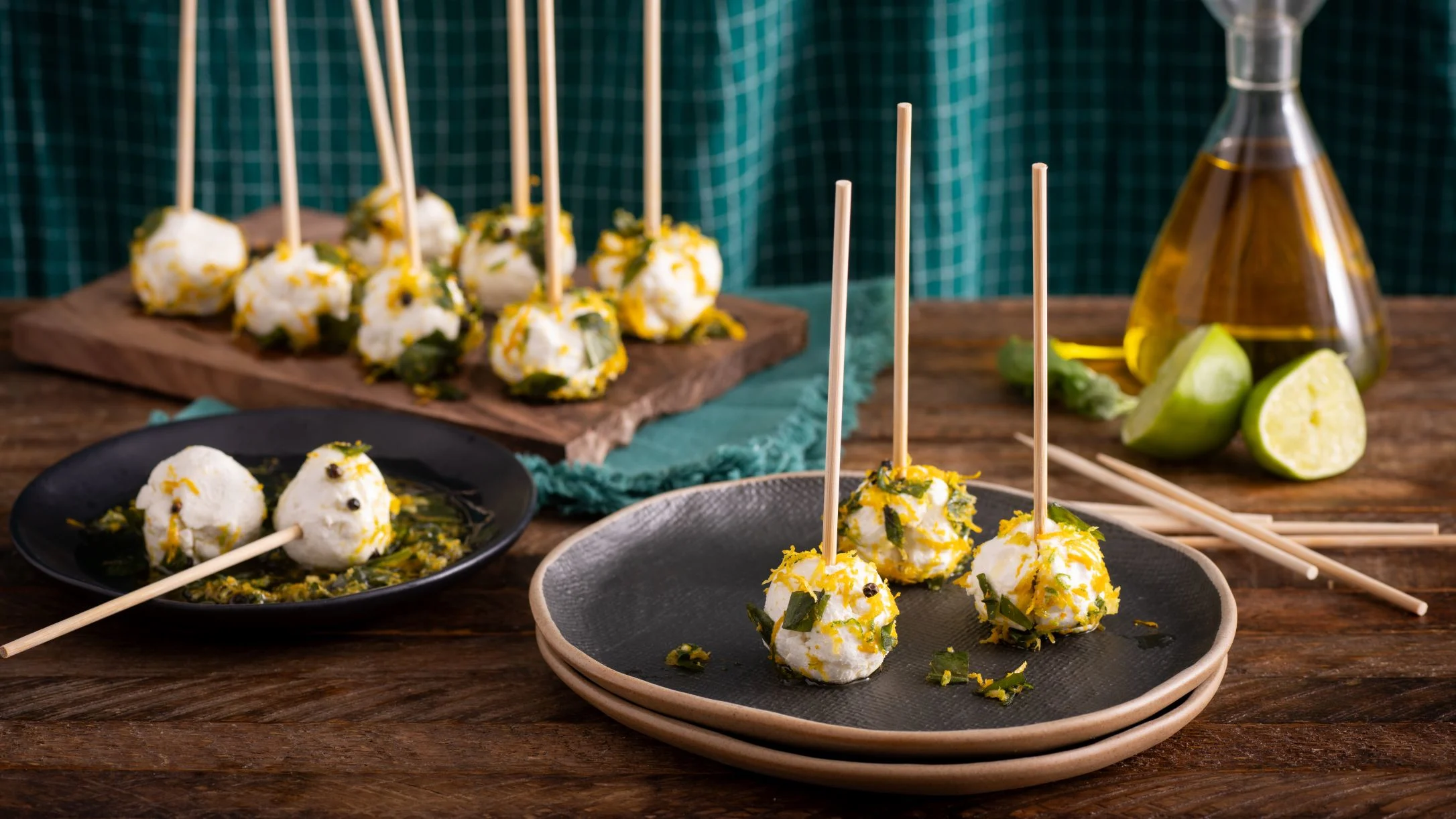 Cheese appetizers on sticks garnished with herbs and citrus zest, served on plates with lime and olive oil nearby.