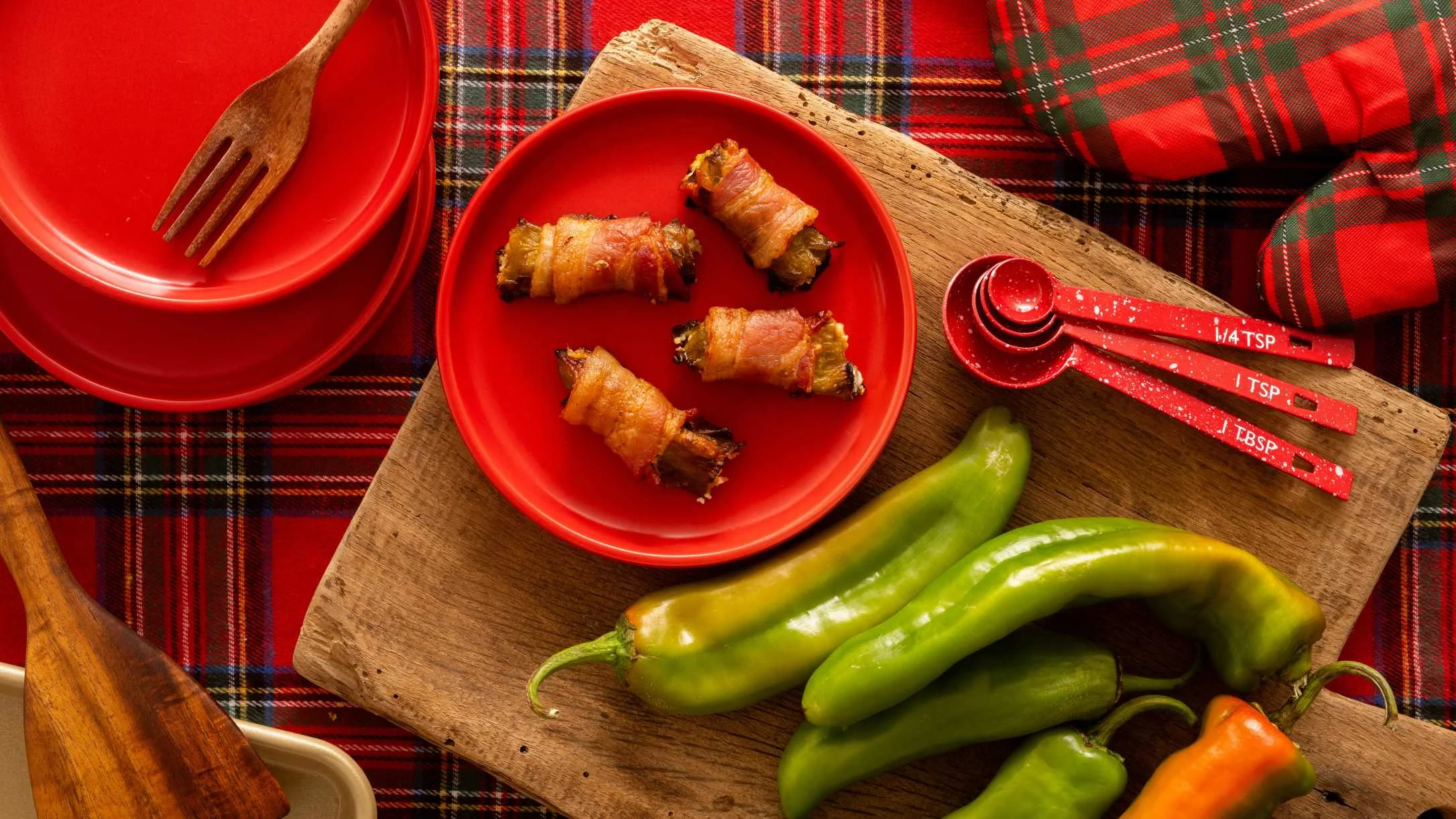 Bacon-wrapped appetizers in red bowl with green peppers on wooden board, red measuring spoons, plaid tablecloth.