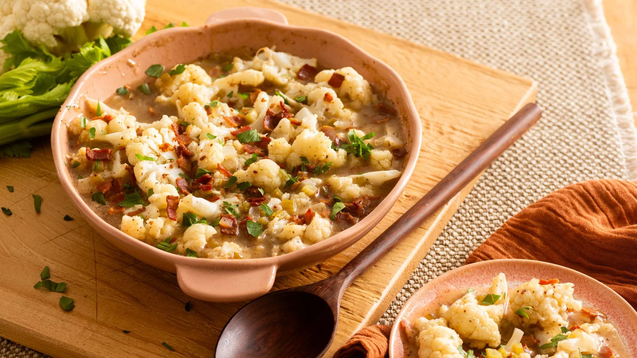 Cauliflower with bacon bits and herbs in a pink bowl, served with wooden spoon on cutting board.