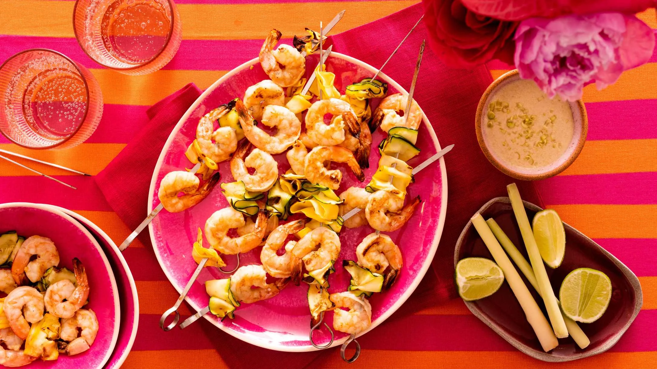 Grilled shrimp skewers with vegetables on a pink plate, served with dipping sauce, lime wedges, and drinks on a colorful tablecloth.