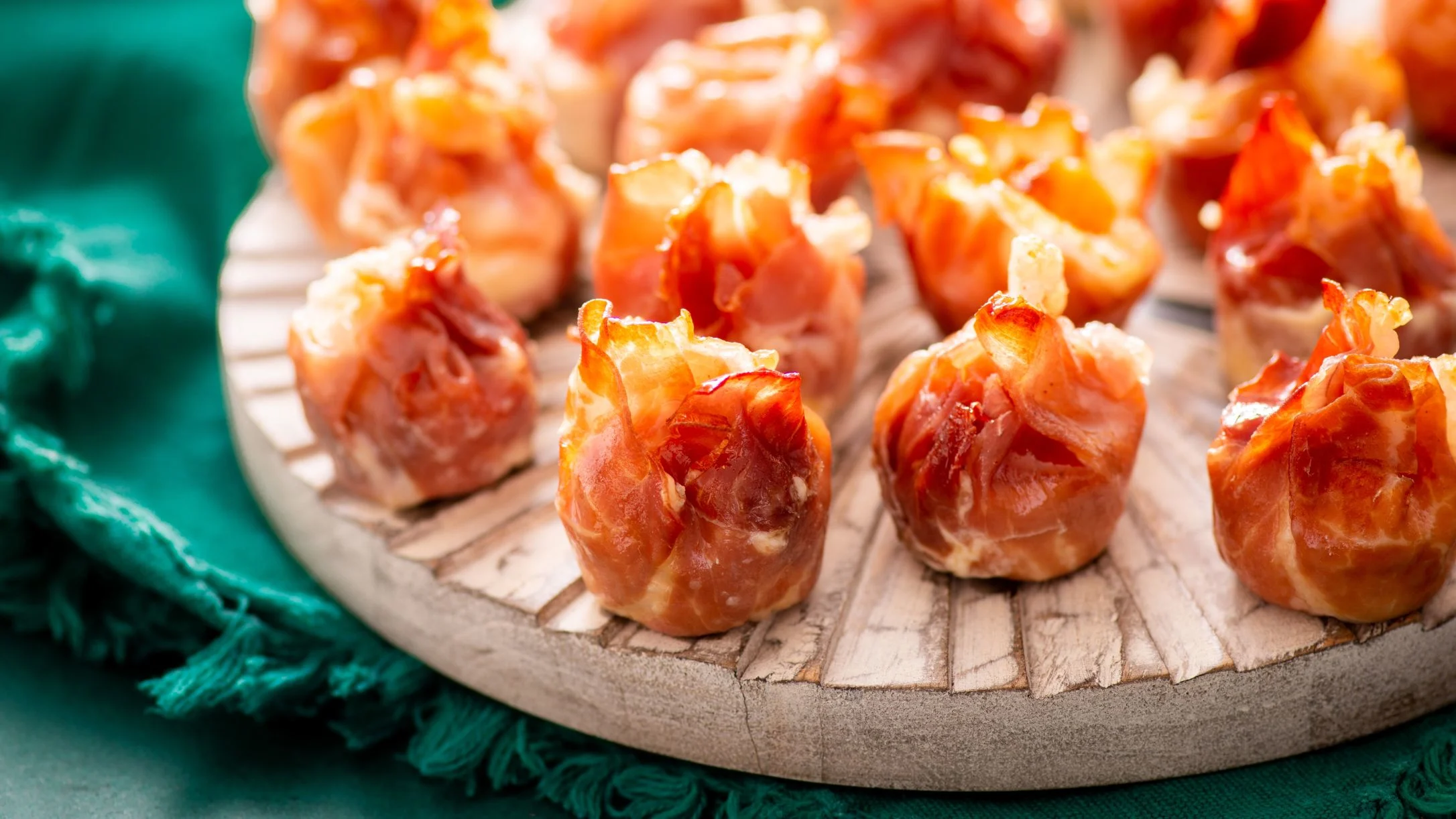 Crispy prosciutto-wrapped appetizers arranged on a wooden serving board against a teal green background.