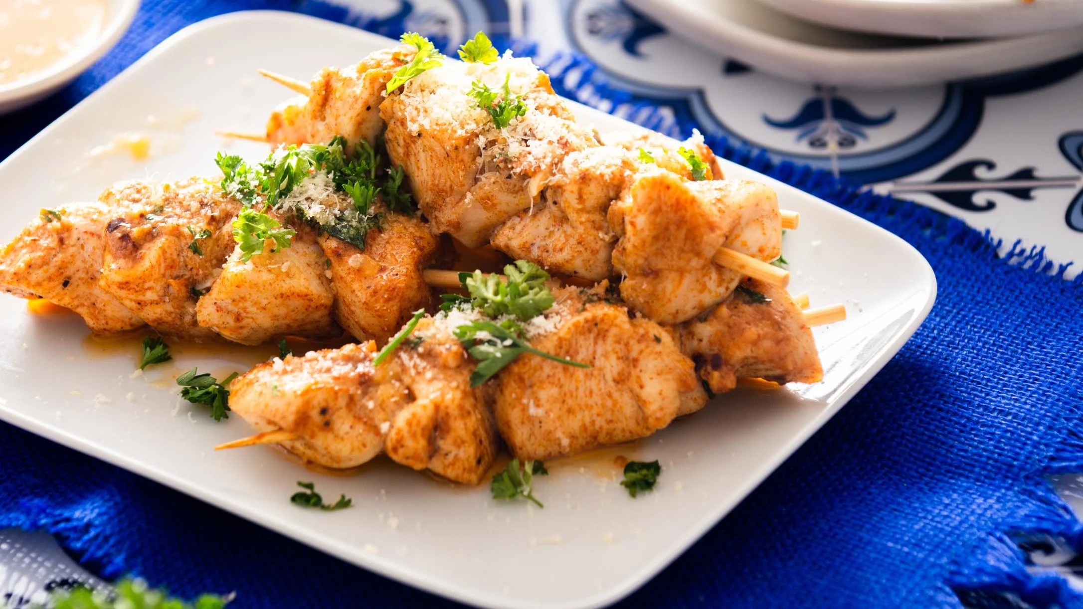 Grilled chicken skewers on white plate, garnished with fresh herbs and grated cheese, served on blue patterned tablecloth.