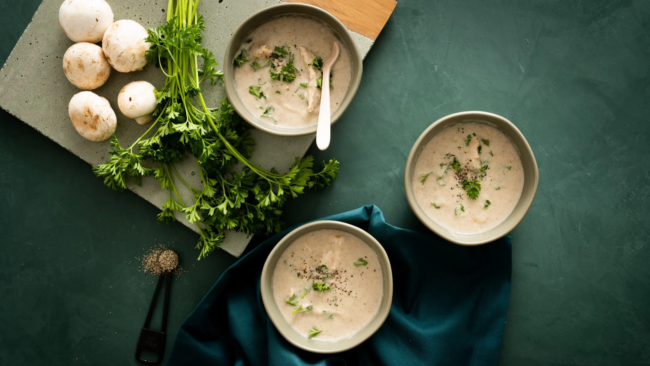 Three bowls of creamy mushroom soup garnished with parsley, with fresh mushrooms and herbs on a green background.