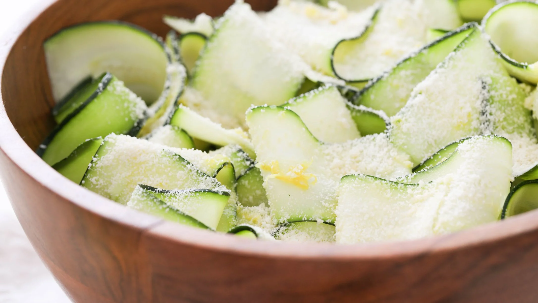 Close up of zucchini ribbons dressed with cheese and lemon zest in a wooden bowl.