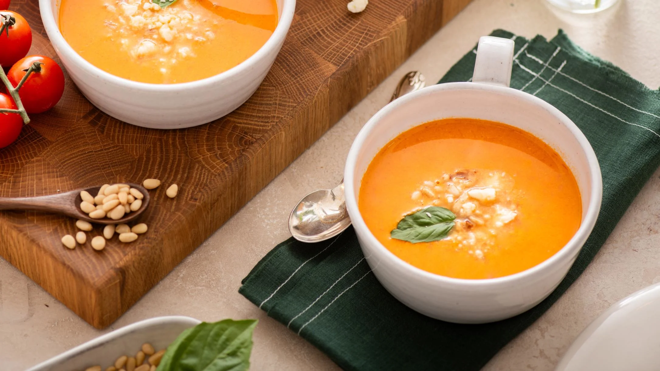 Creamy orange tomato soup in white mugs garnished with basil, served with pine nuts and cherry tomatoes on wooden board.