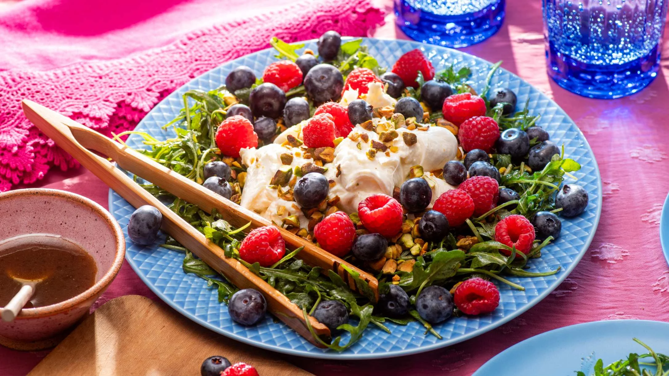 Fresh salad with burrata cheese, blueberries, raspberries, and arugula on a blue plate with wooden serving utensils.