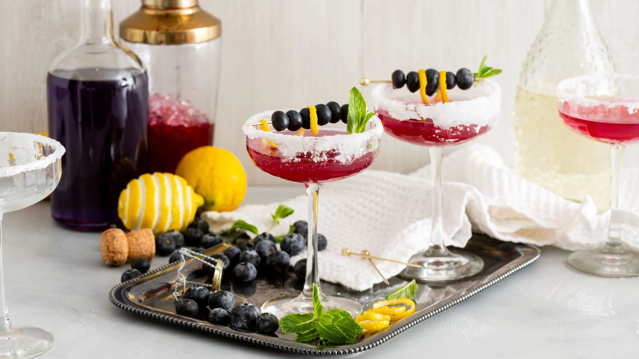 Red cocktails with sugar-rimmed glasses garnished with blueberries and lemon, served on a silver tray with fresh ingredients.