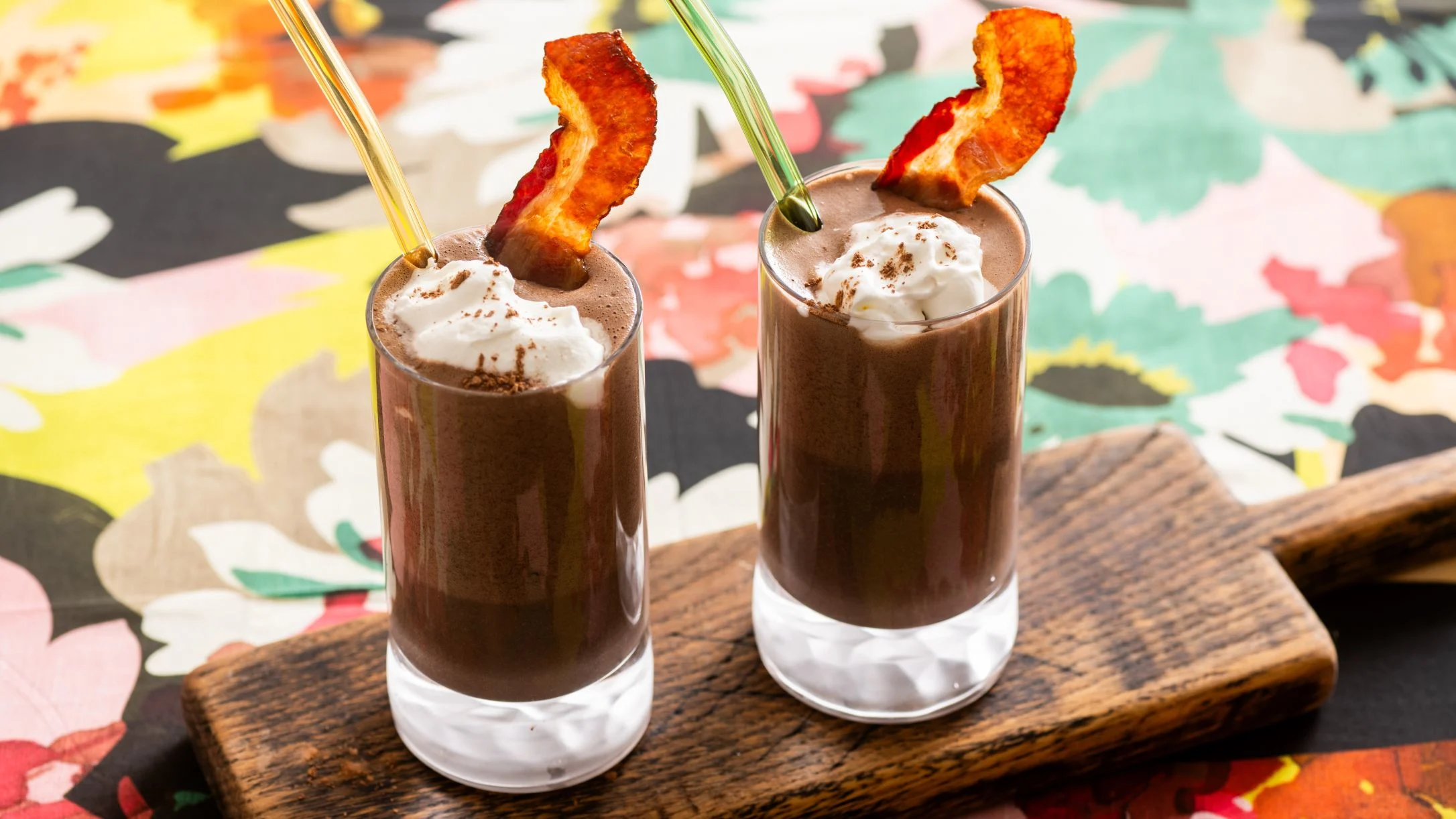 Two chocolate cocktails in small glasses topped with whipped cream and crispy bacon garnish on a wooden board.