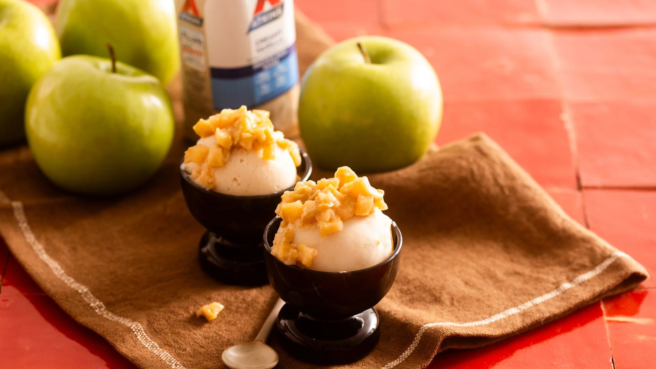 Vanilla ice cream topped with apple pieces in dark egg cups, with green apples and shake bottle in background.