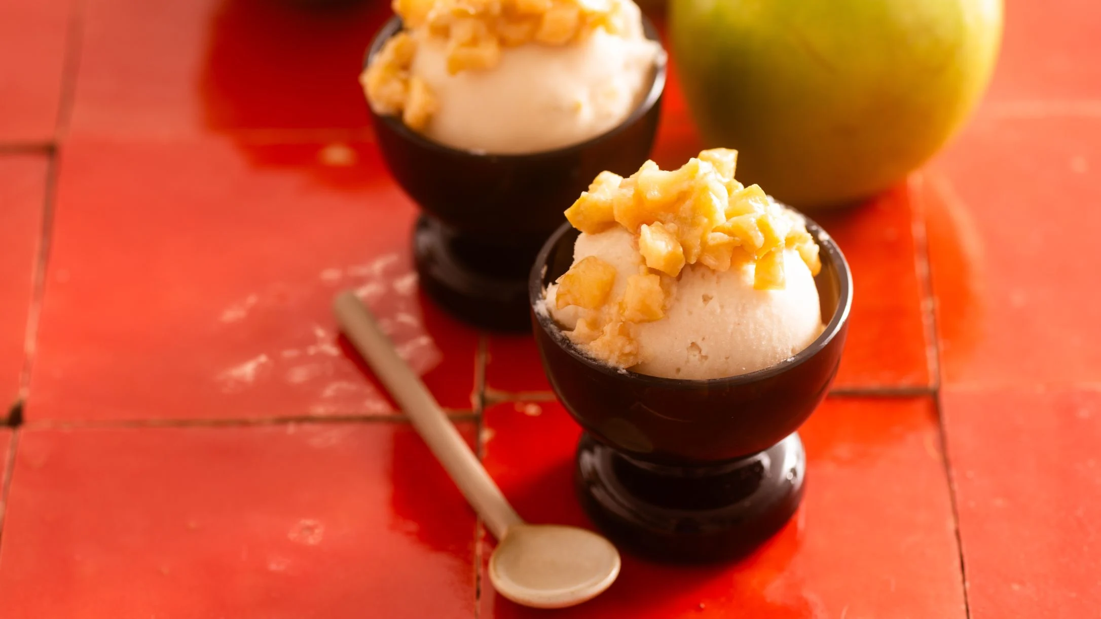 Two dark cups of vanilla ice cream topped with diced apple pieces on a bright red surface, with a spoon and whole apple nearby.