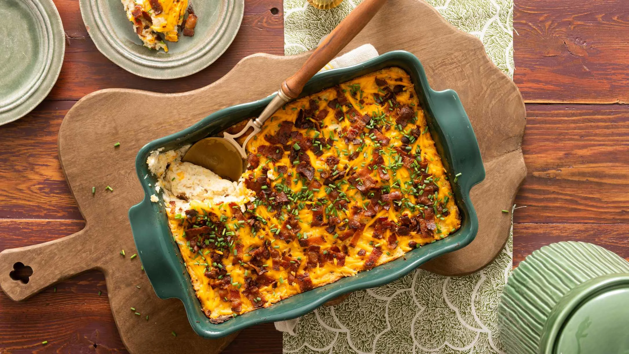 Green baking dish with cheesy faux potato casserole topped with bacon and chives, served with a gold spoon on wooden board.