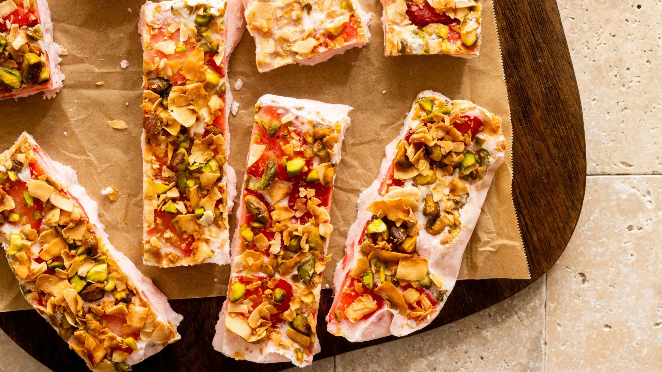 Sliced frozen yogurt topped with nuts, pistachios, and strawberries arranged on brown parchment paper on a wooden board.