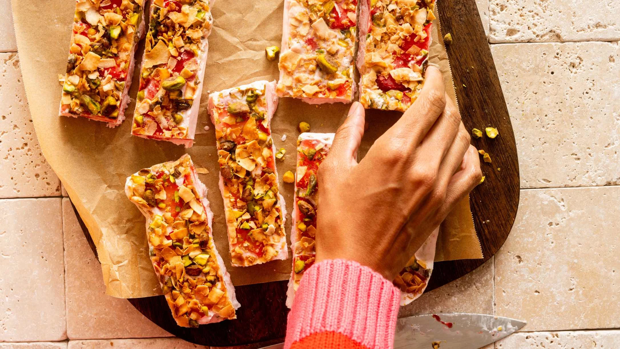 Hand in pink sleeve reaching for colorful dessert bars topped with nuts and fruits on wooden board with parchment paper.