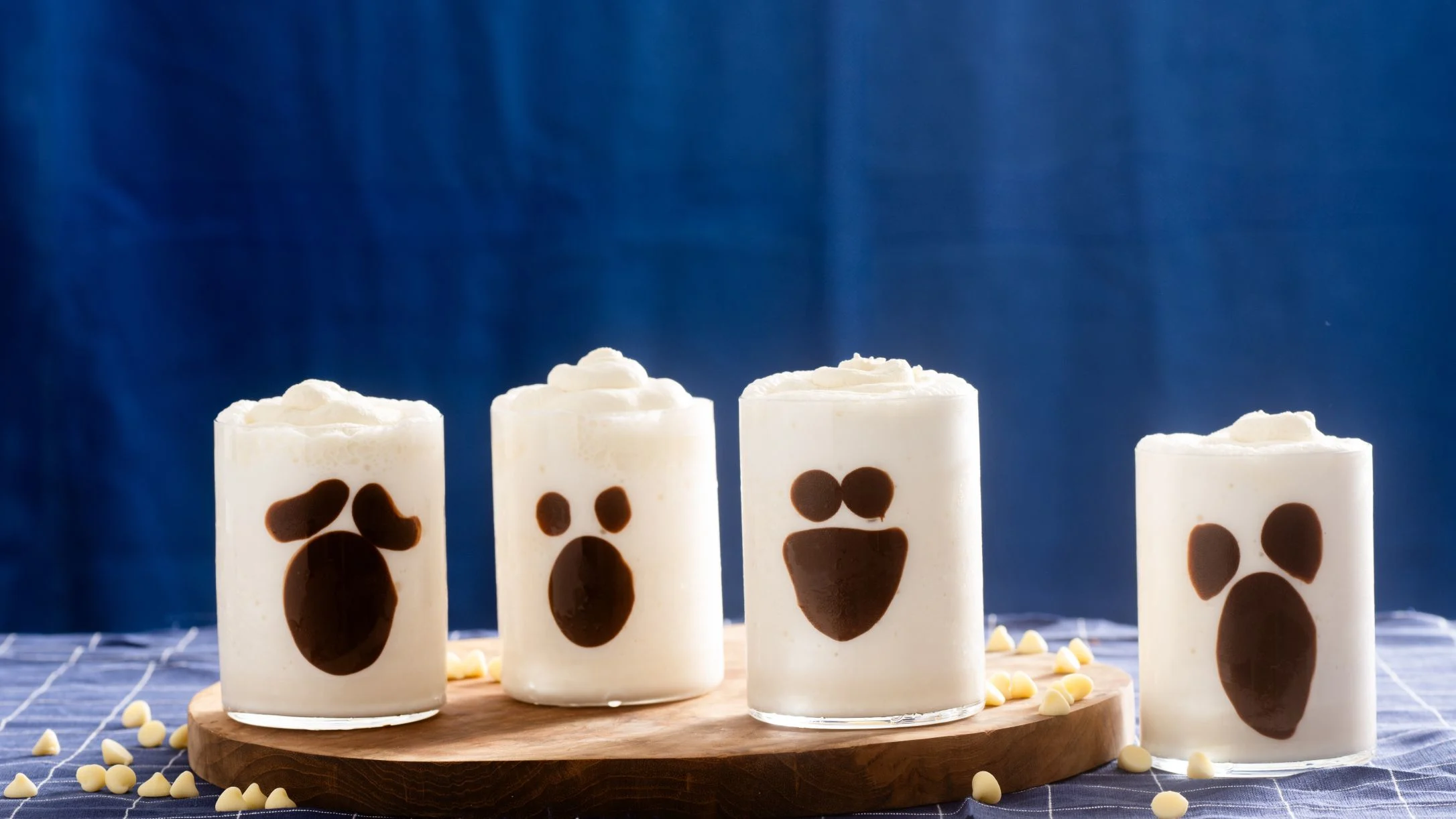 Four white dessert cocktails with chocolate ghost face designs arranged on a wooden board against a blue background.