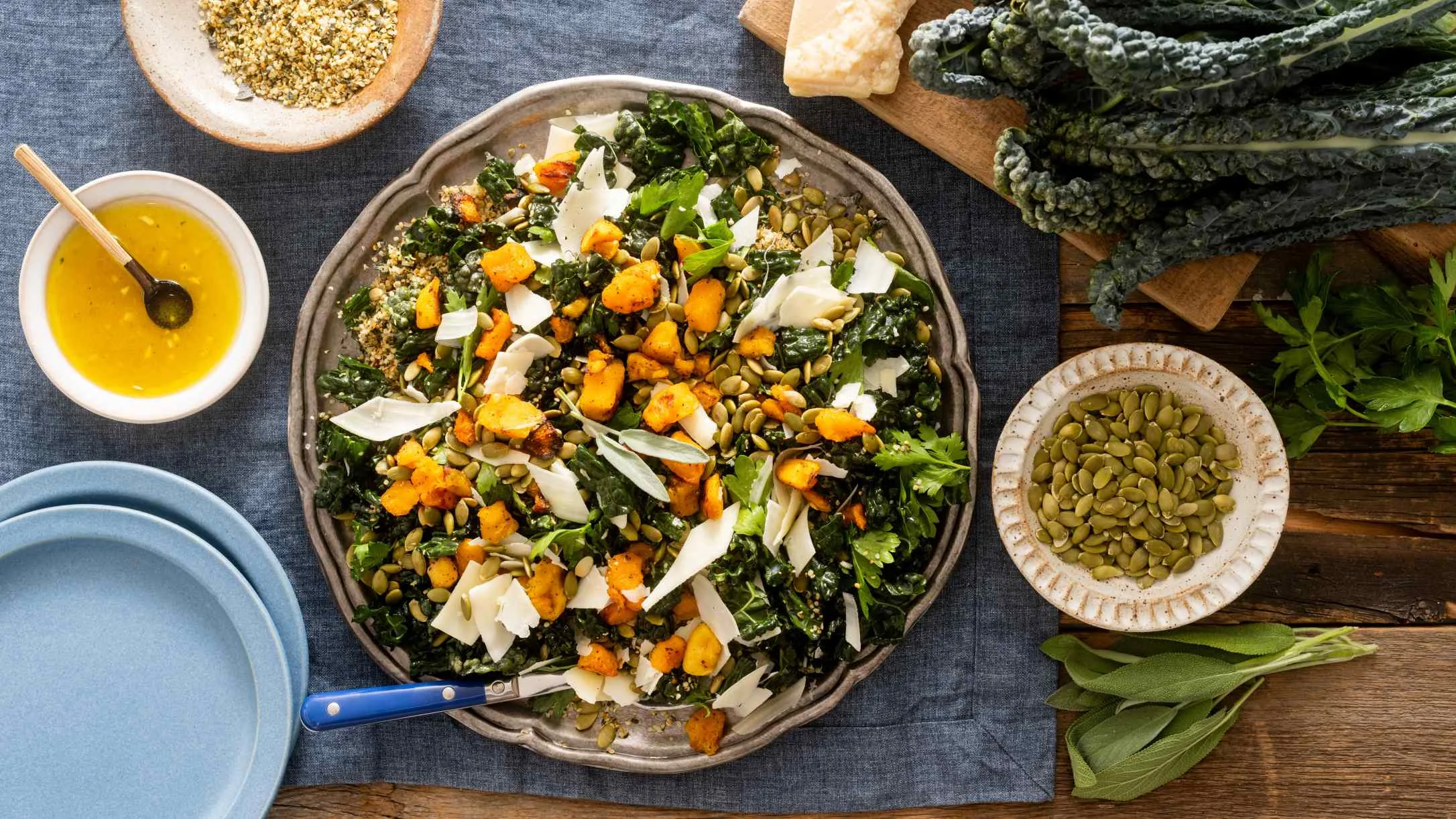 Kale salad with roasted butternut squash, cheese shavings, and pumpkin seeds, surrounded by ingredients and dressing.