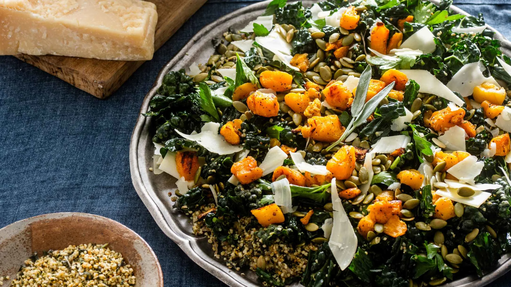 Kale salad with roasted butternut squash, parmesan shavings, and pumpkin seeds, with cheese block and hemp seeds bowl nearby.