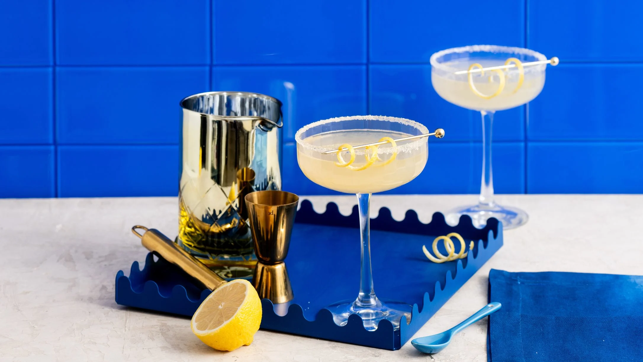 Two cocktails with sugar rims and lemon twists on a blue tray with bartending tools and half a lemon.