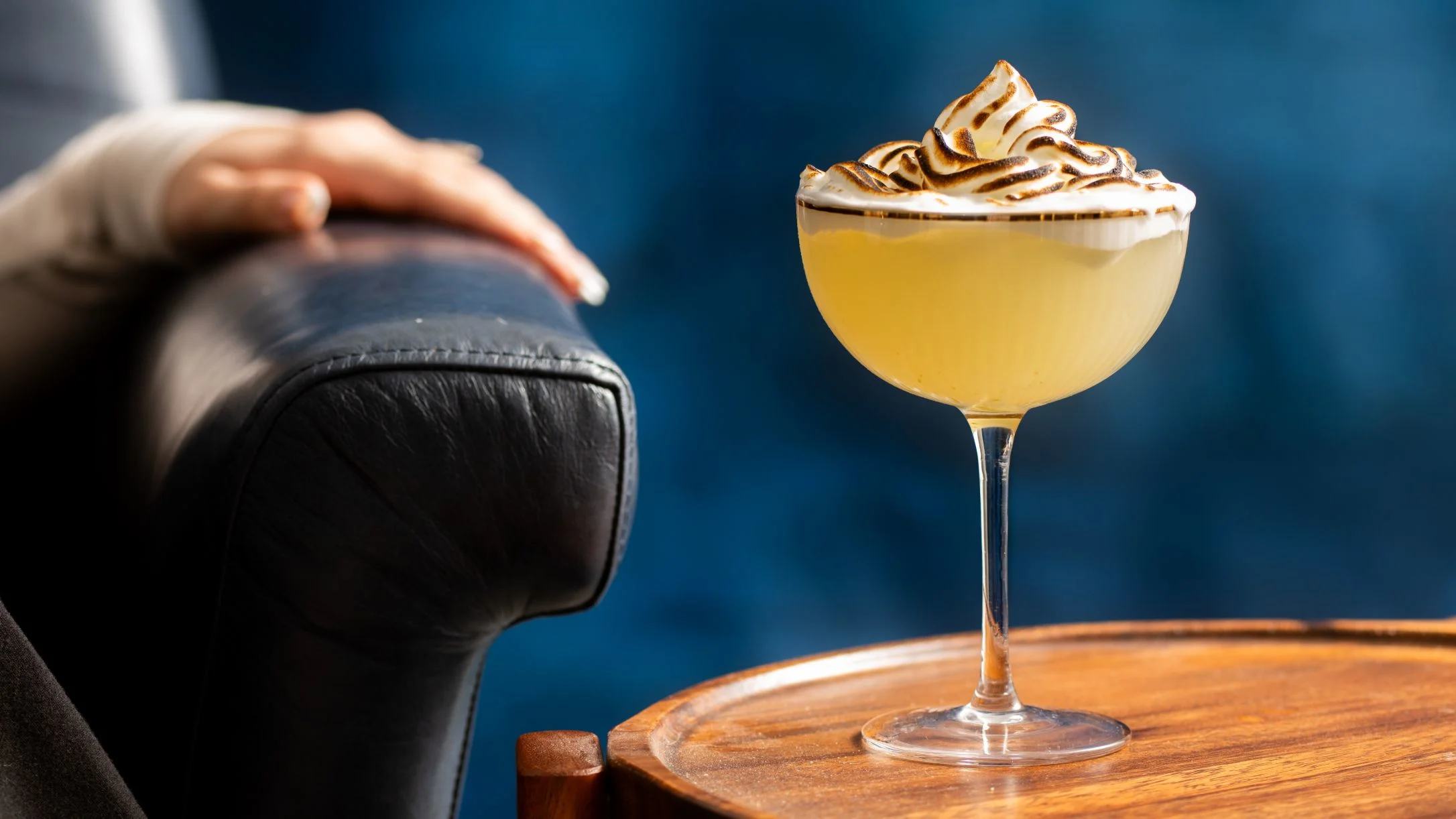 Yellow cocktail with toasted meringue topping on wooden table beside leather chair against blue background.