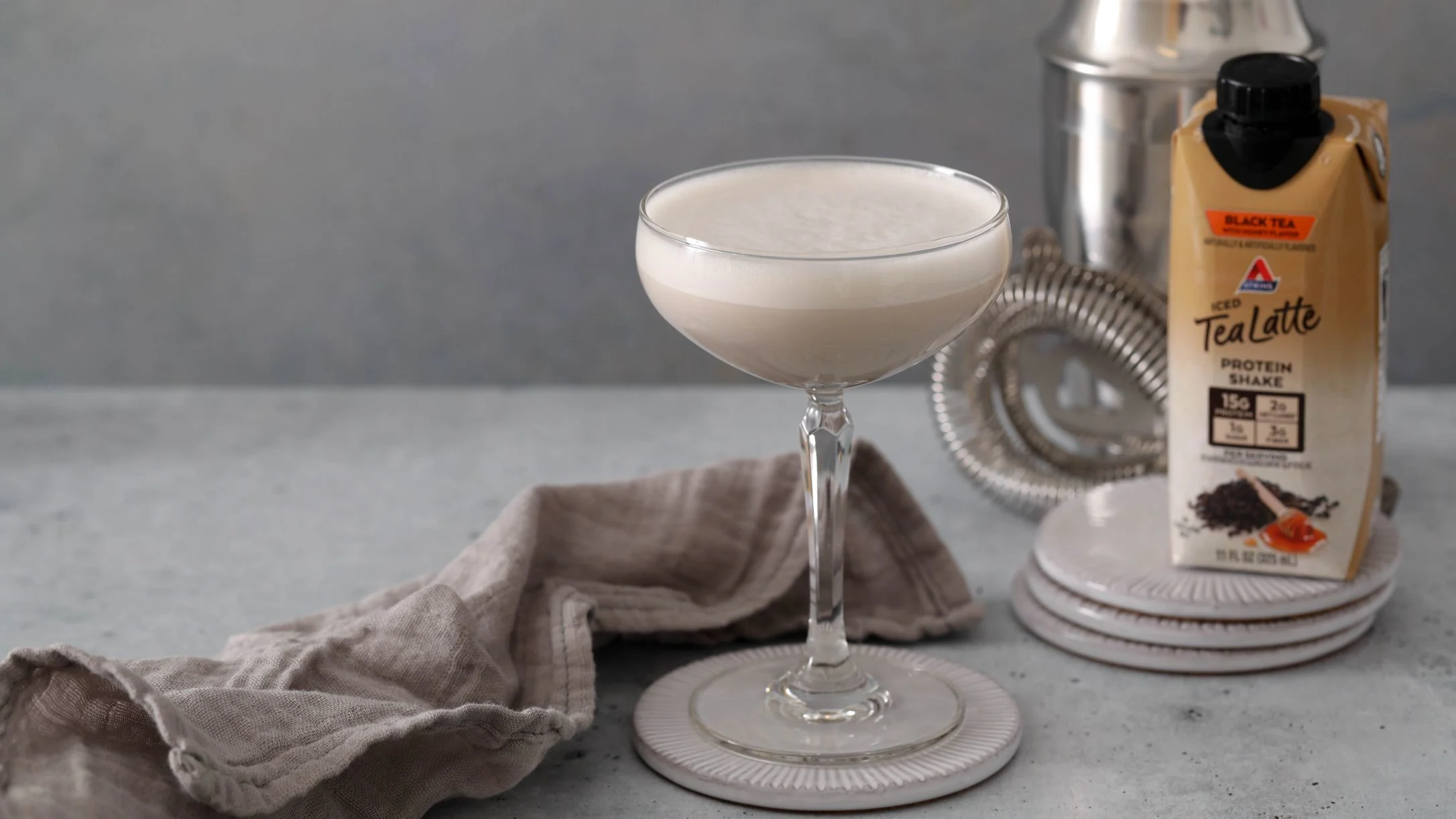 Creamy cocktail in coupe glass with TeaLatte protein shake carton, cocktail shaker, and linen napkin on marble surface.