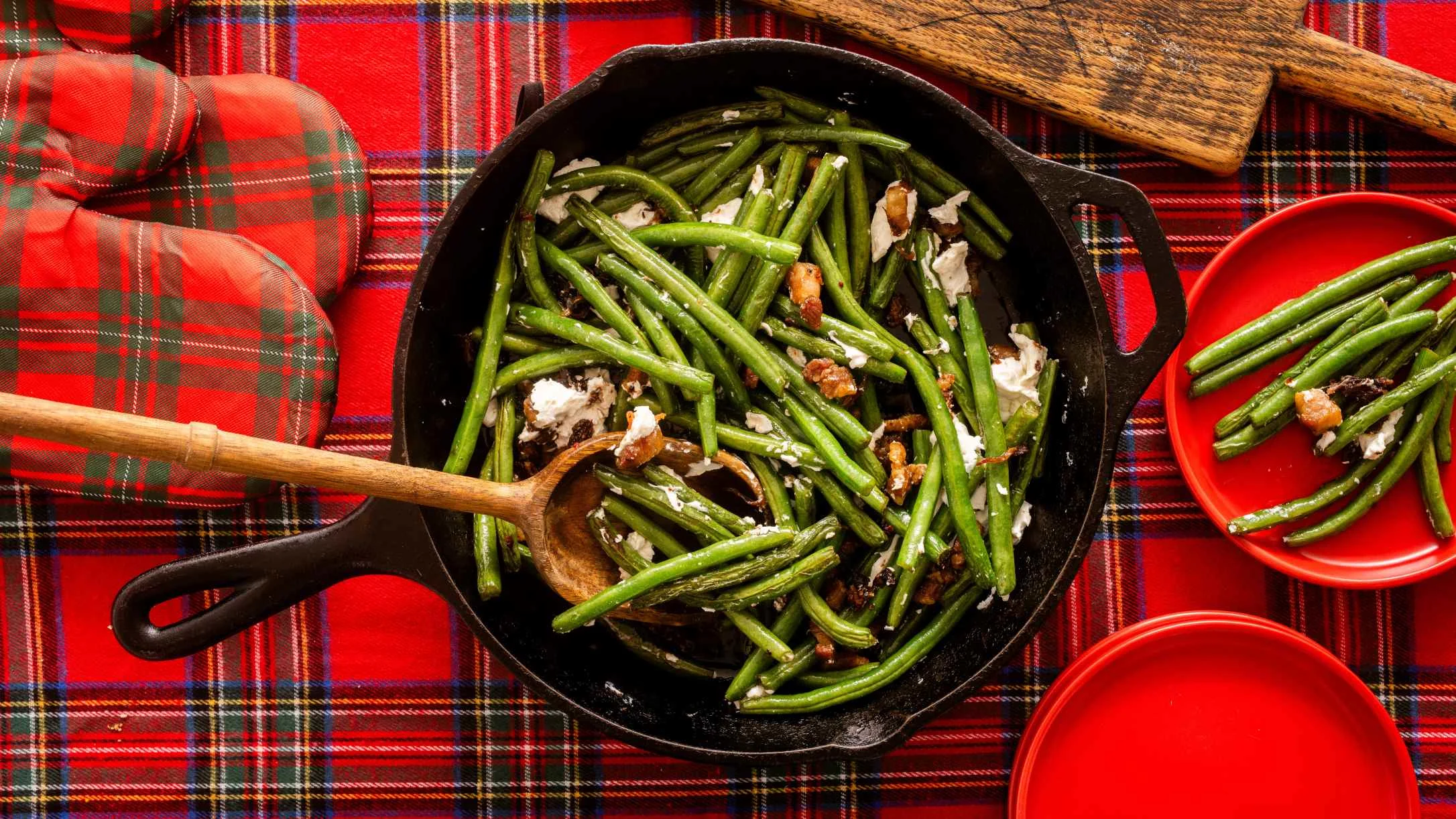 Cast iron skillet with sautéed green beans and cheese on a red plaid tablecloth, wooden spoon and red plates nearby.