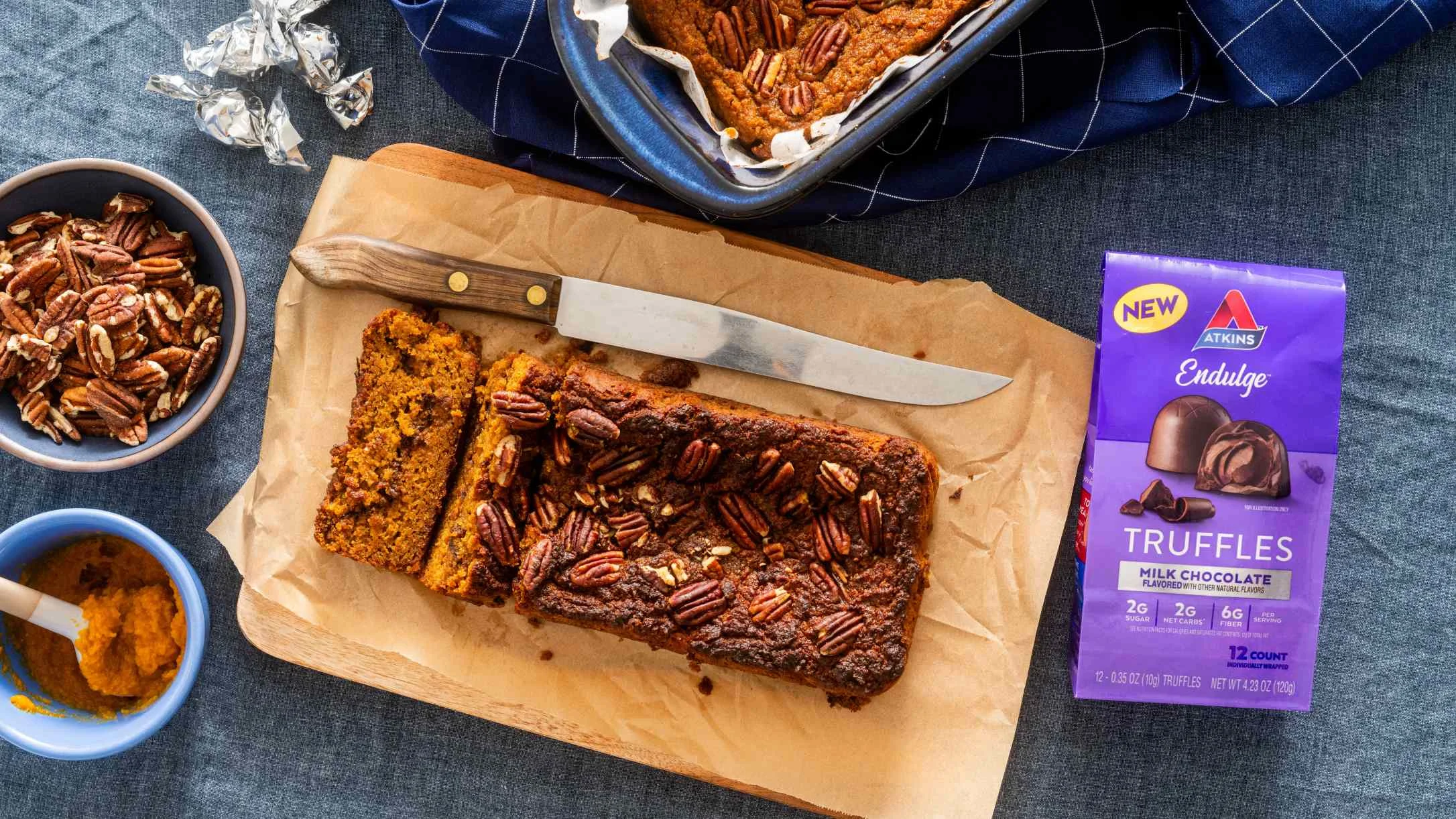 Sliced pumpkin bread with pecans on parchment paper, alongside Atkins chocolate truffles and bowls of ingredients.