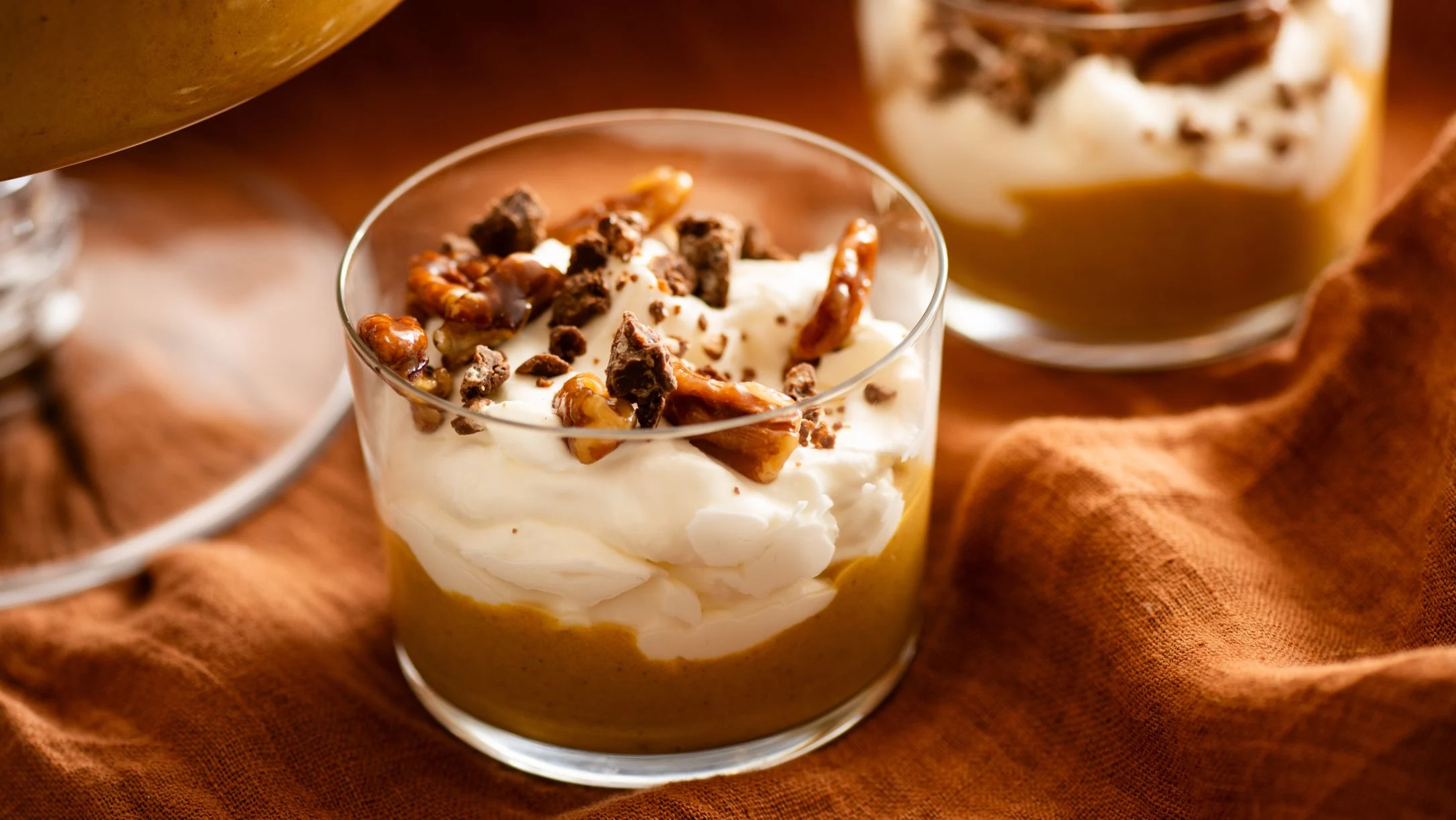 Layered pumpkin dessert in glass with whipped cream, chocolate pieces and nuts on orange fabric background.