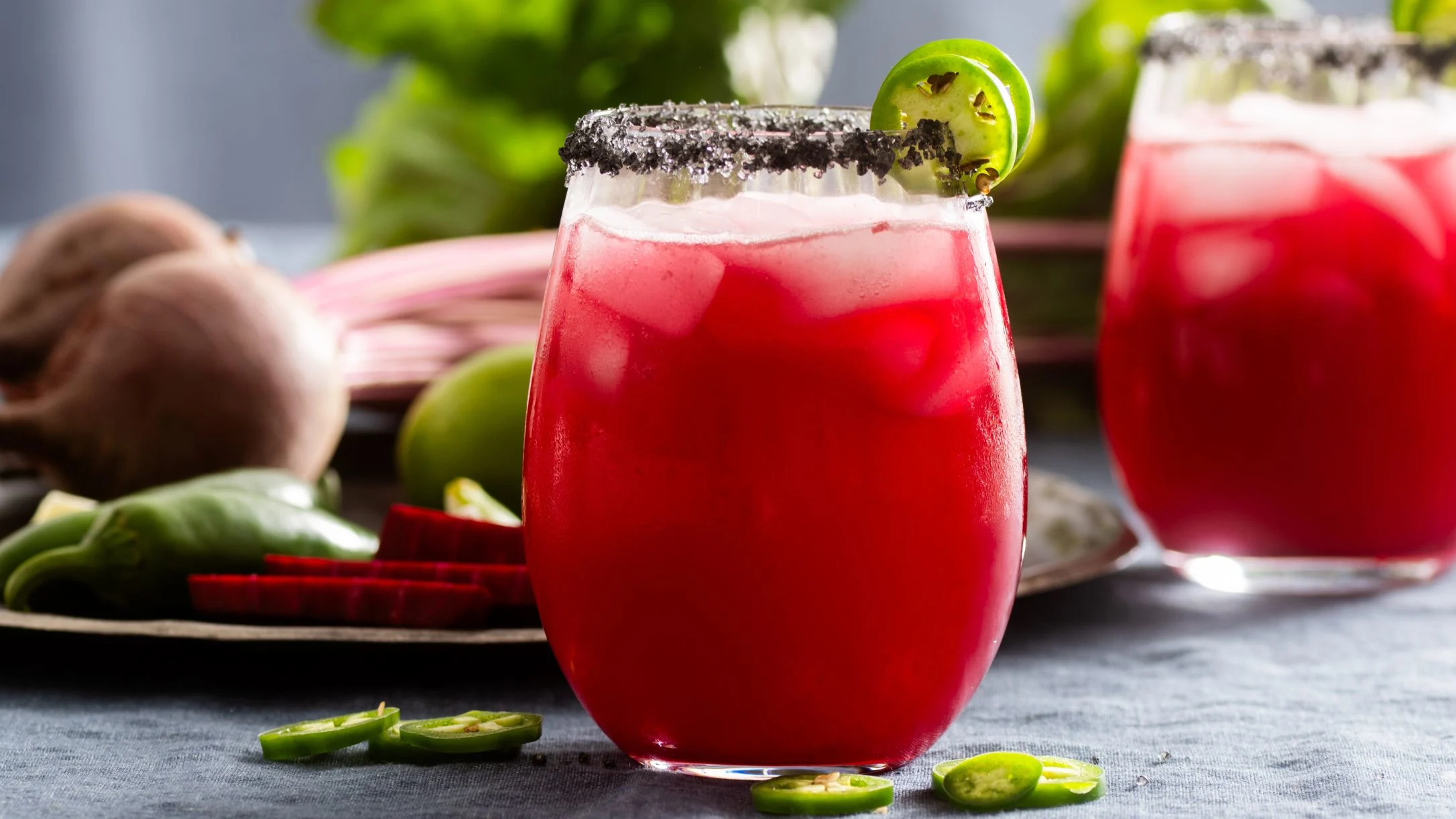 Vibrant red cocktail with salt rim and lime garnish, served with fresh beets and jalapeño slices nearby.