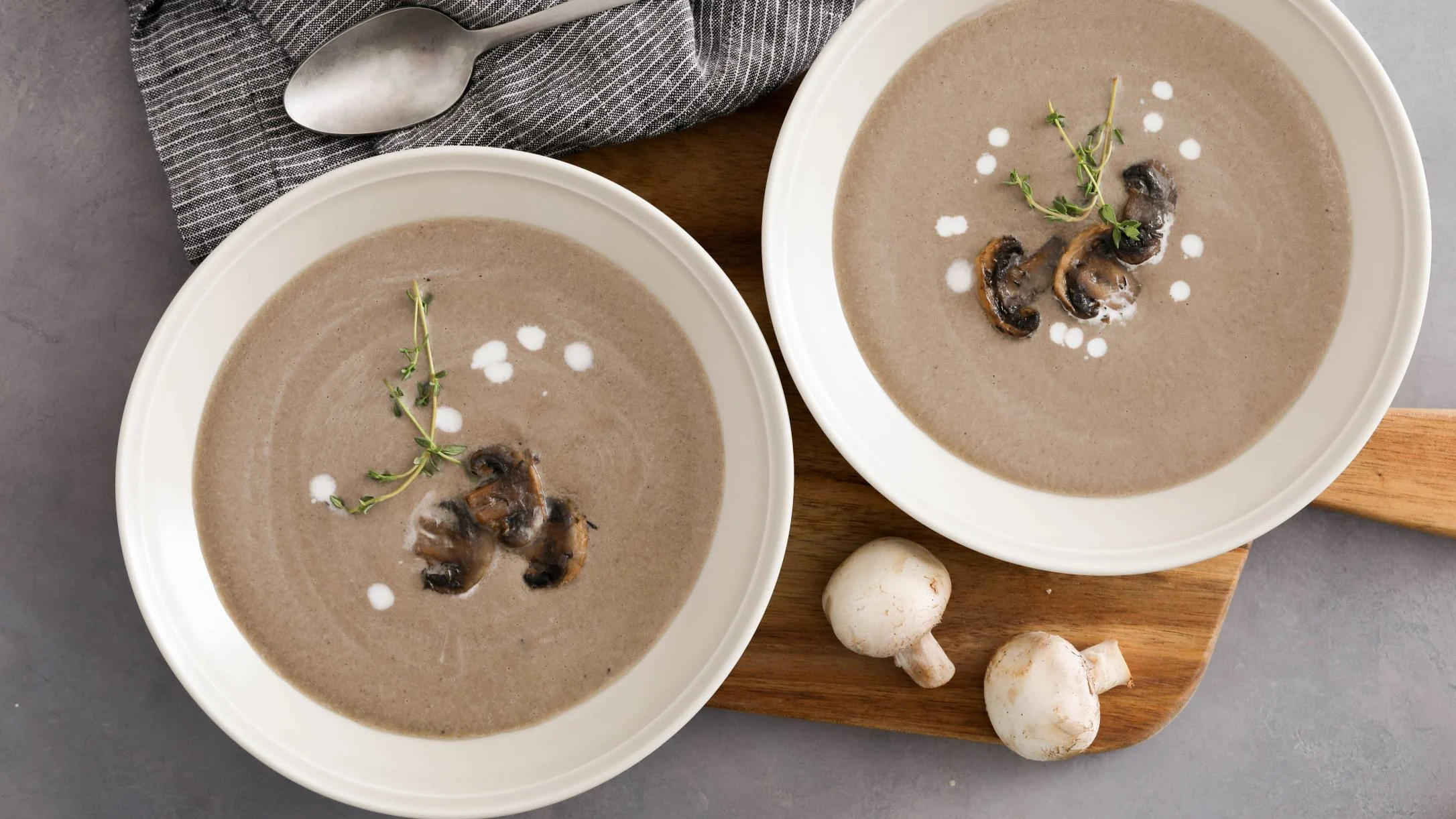 Two white bowls of creamy mushroom soup garnished with thyme and cream dots, served with fresh mushrooms on a wooden board.