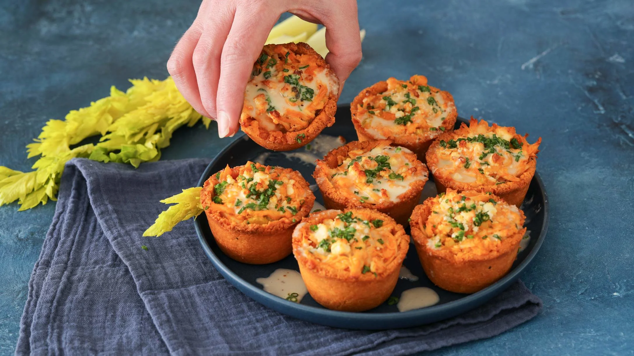 Hand picking up a savory muffin from a plate of six crispy, cheese-topped appetizers garnished with herbs.