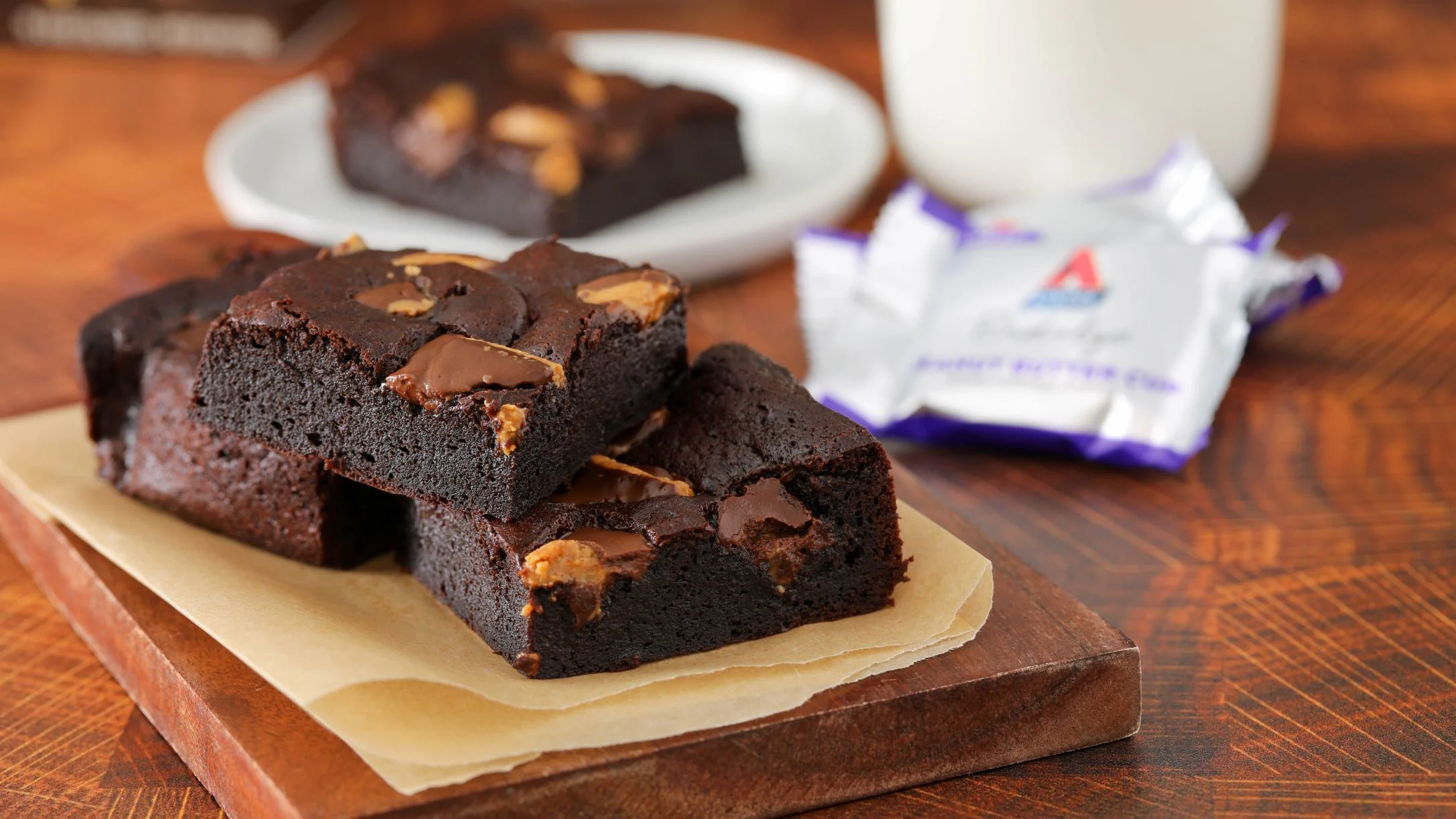 Rich chocolate brownies with nuts on wooden board, with more brownies on plate and candy wrapper in background.