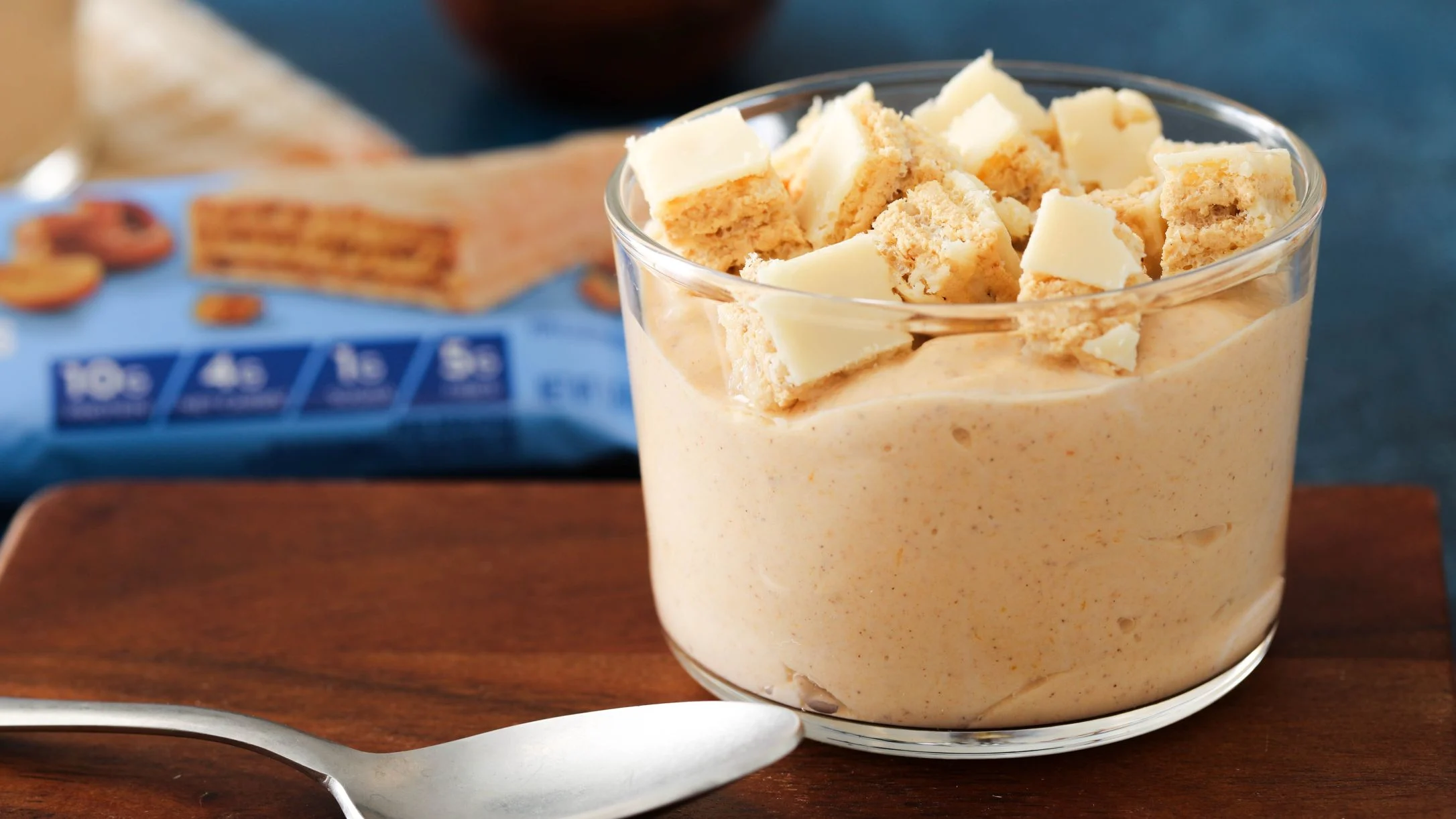 Creamy yogurt in glass cup topped with wafer bar pieces, with protein bar packaging in background.