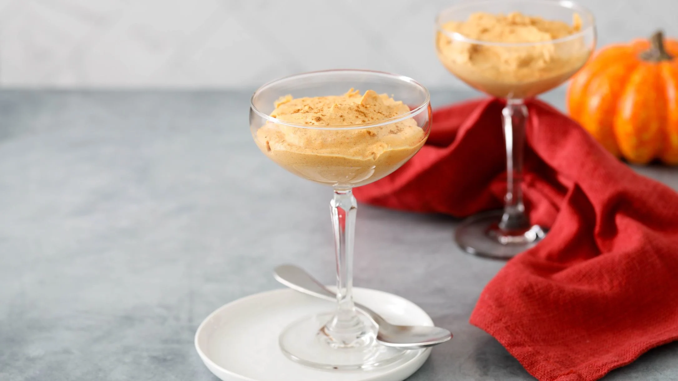 Two glasses of pumpkin mousse dessert with a small orange pumpkin and red napkin on a gray marble surface.
