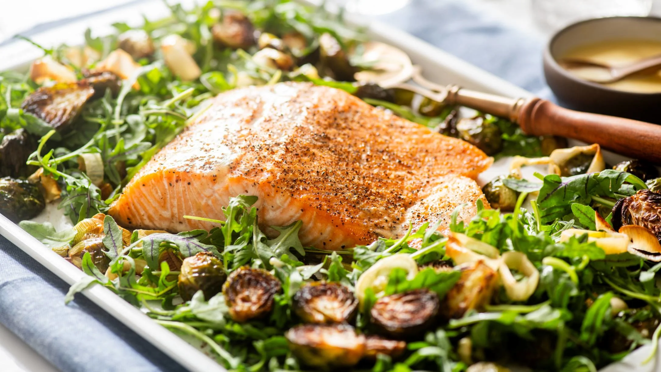 Seasoned salmon fillet on a baking sheet with roasted vegetables and fresh greens, sauce bowl visible nearby.
