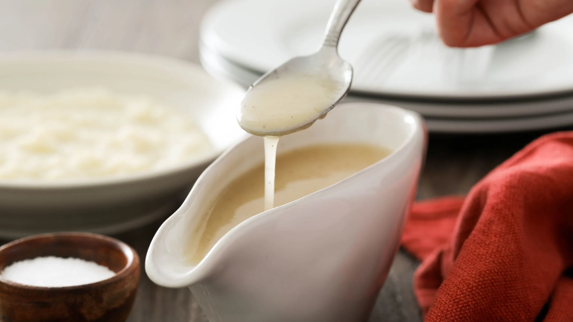 Creamy sauce being poured from a spoon into a white gravy boat, with a salt dish and plates in the background.
