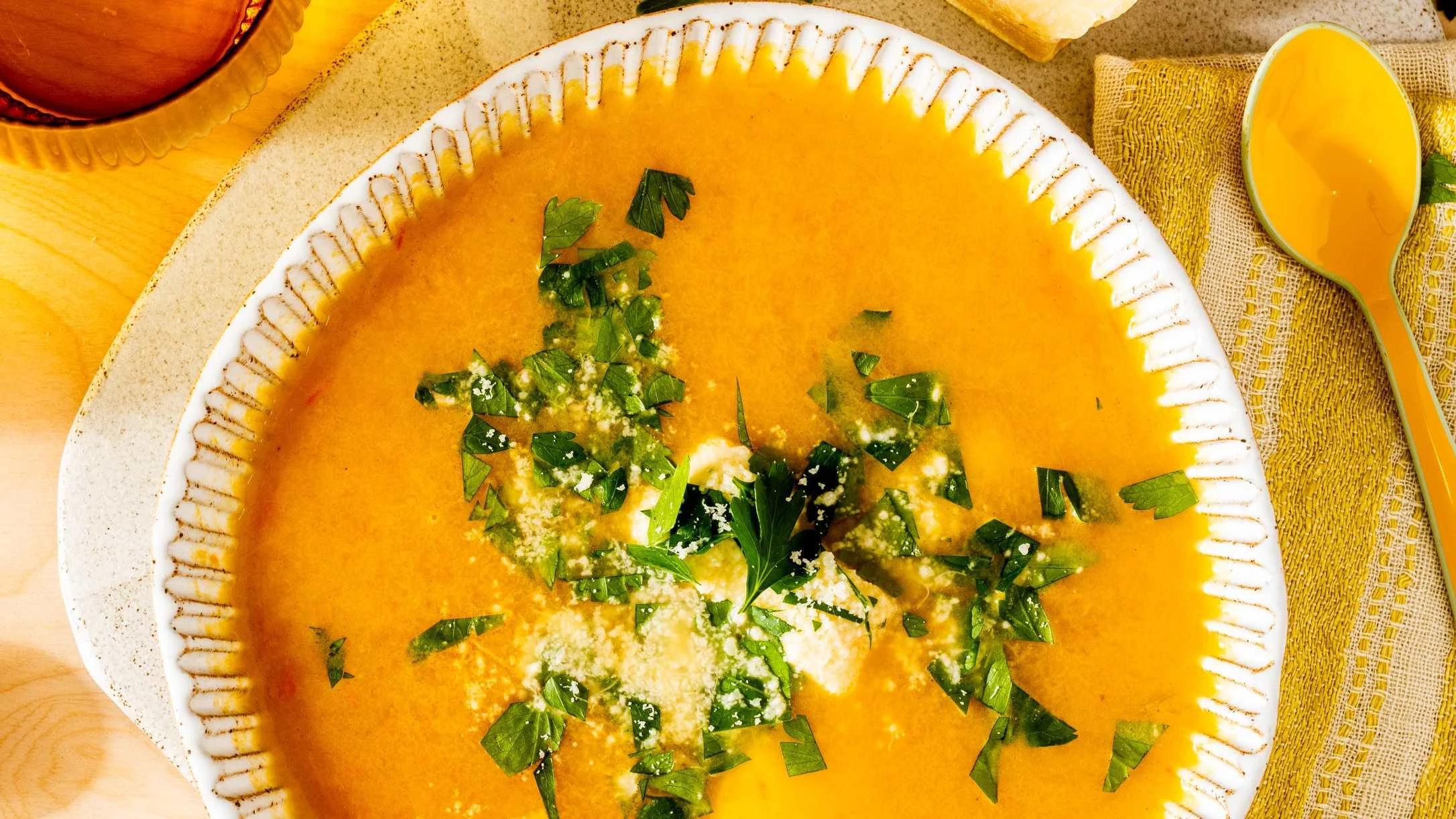 Creamy orange soup in white bowl garnished with fresh herbs and cheese, served with yellow spoon on burlap.