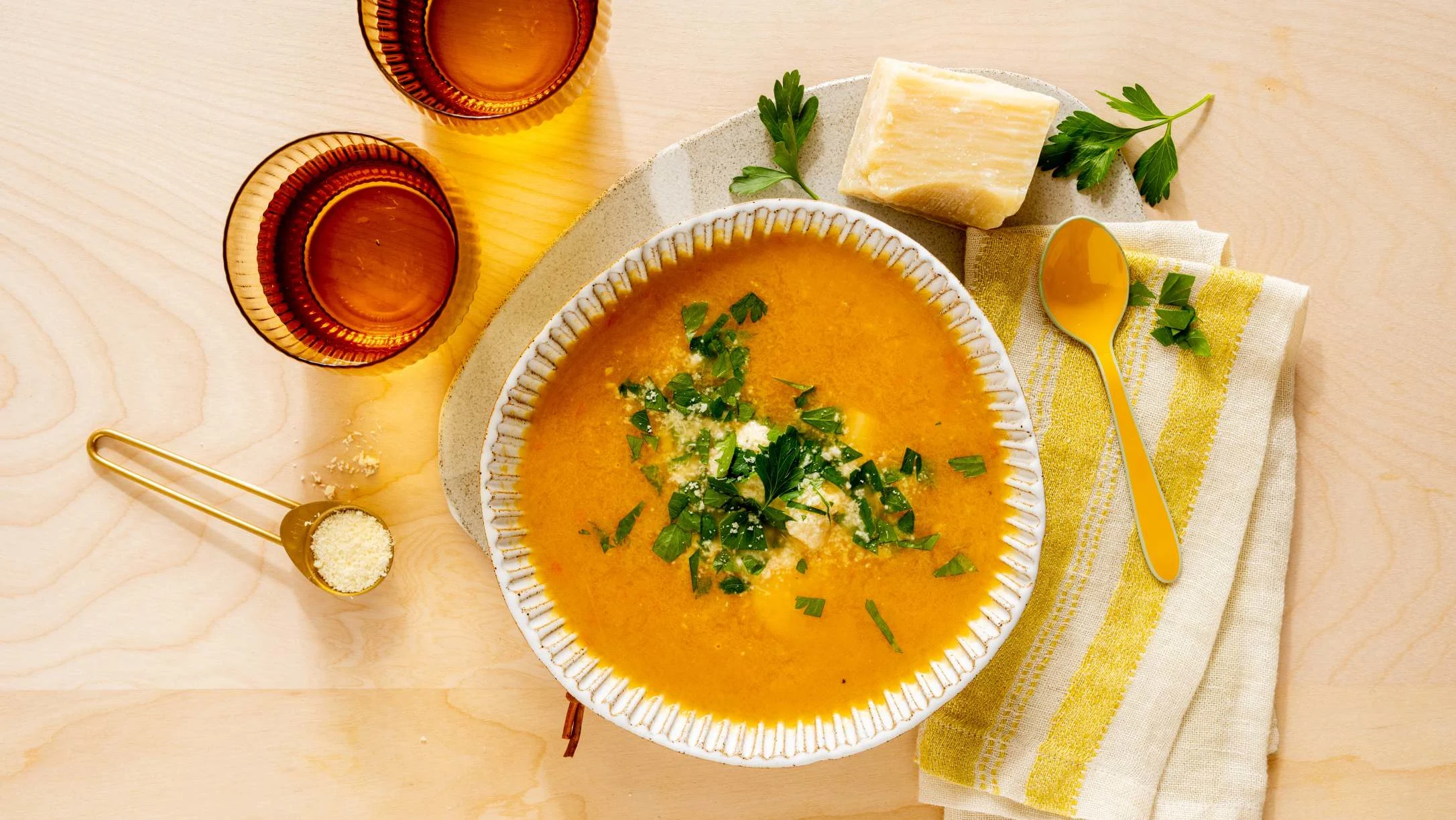 Orange soup garnished with herbs in ceramic bowl, served with parmesan cheese, yellow spoon, and amber glasses on wooden table.