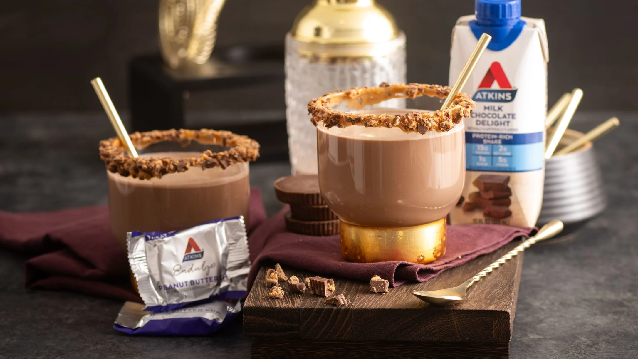 Two chocolate drinks with nut-rimmed glasses on wooden board, alongside Atkins chocolate milk and peanut butter products.