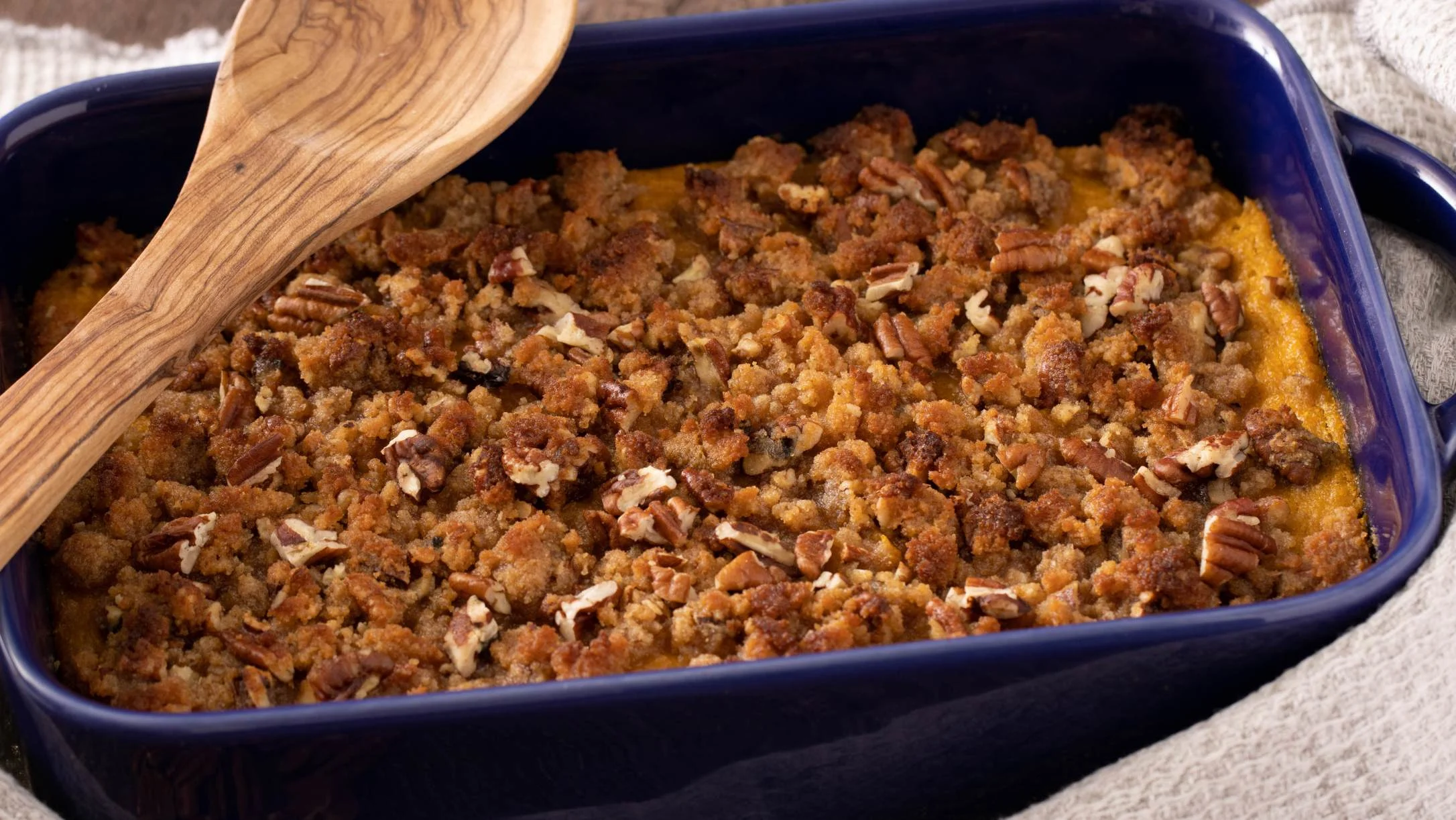 Sweet potato casserole with brown sugar pecan topping in a blue baking dish with wooden spoon.