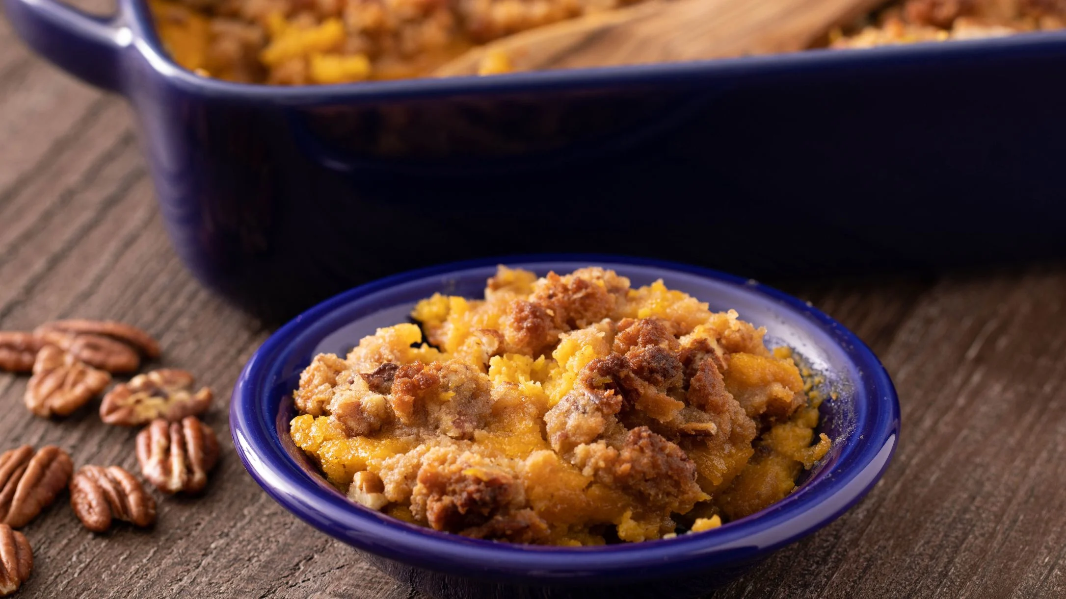Sweet potato casserole with crumbly pecan topping in a blue dish, with baking dish and pecans nearby.