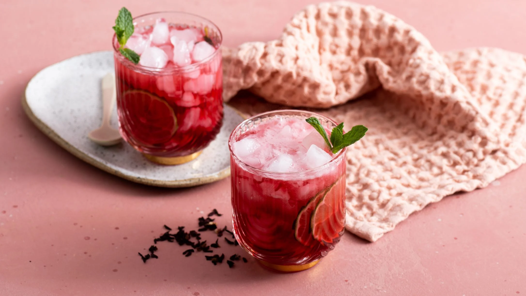 Two vibrant red mocktails with ice and mint garnish on a pink background with textured cloth and tea leaves.